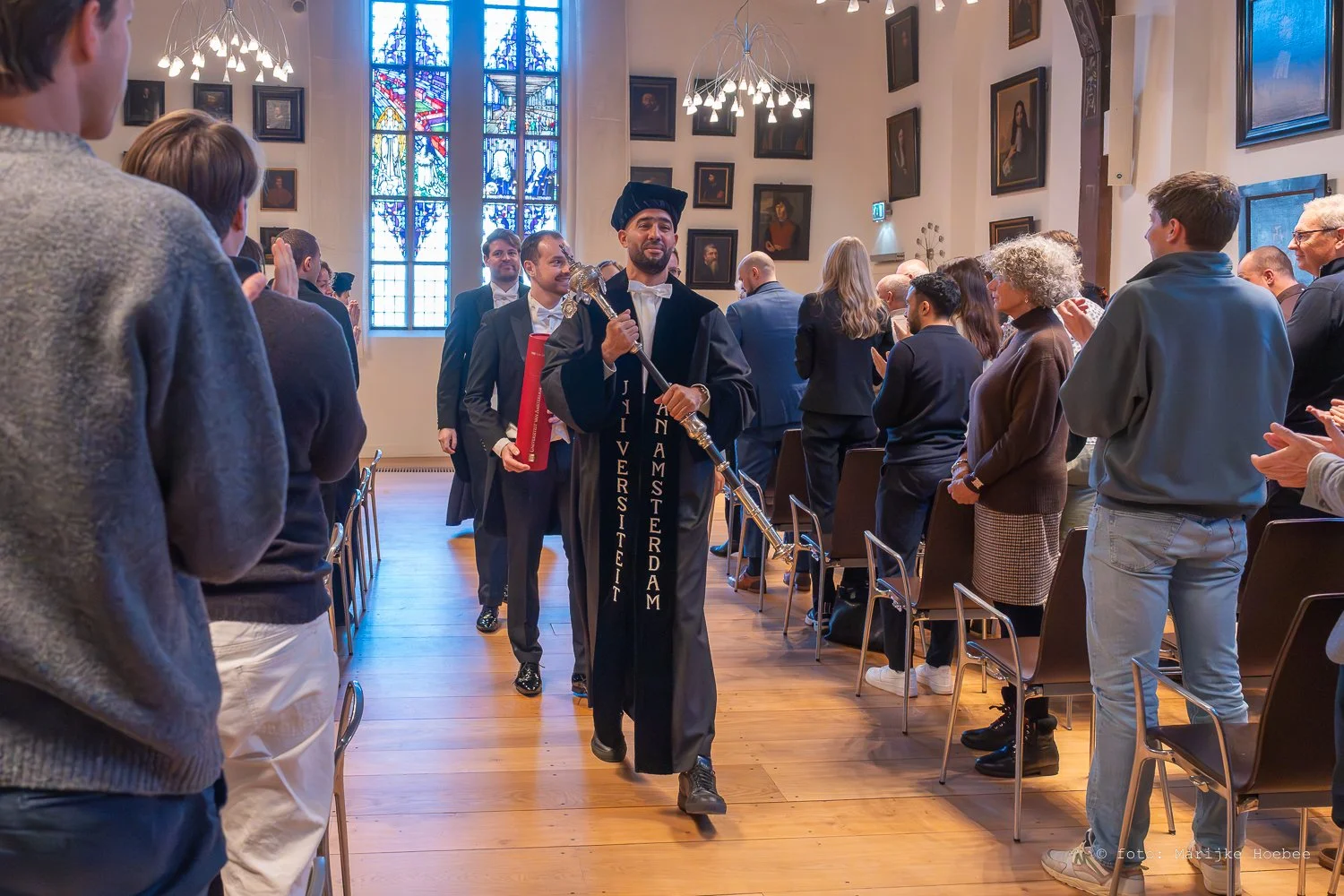 Een ceremoniële gebeurtenis in een kerk met een groep mensen die elkaar applaudisseert en een man in universitaire toga die door de zaal loopt, waarbij de kerkramen en schilderijen zichtbaar zijn.