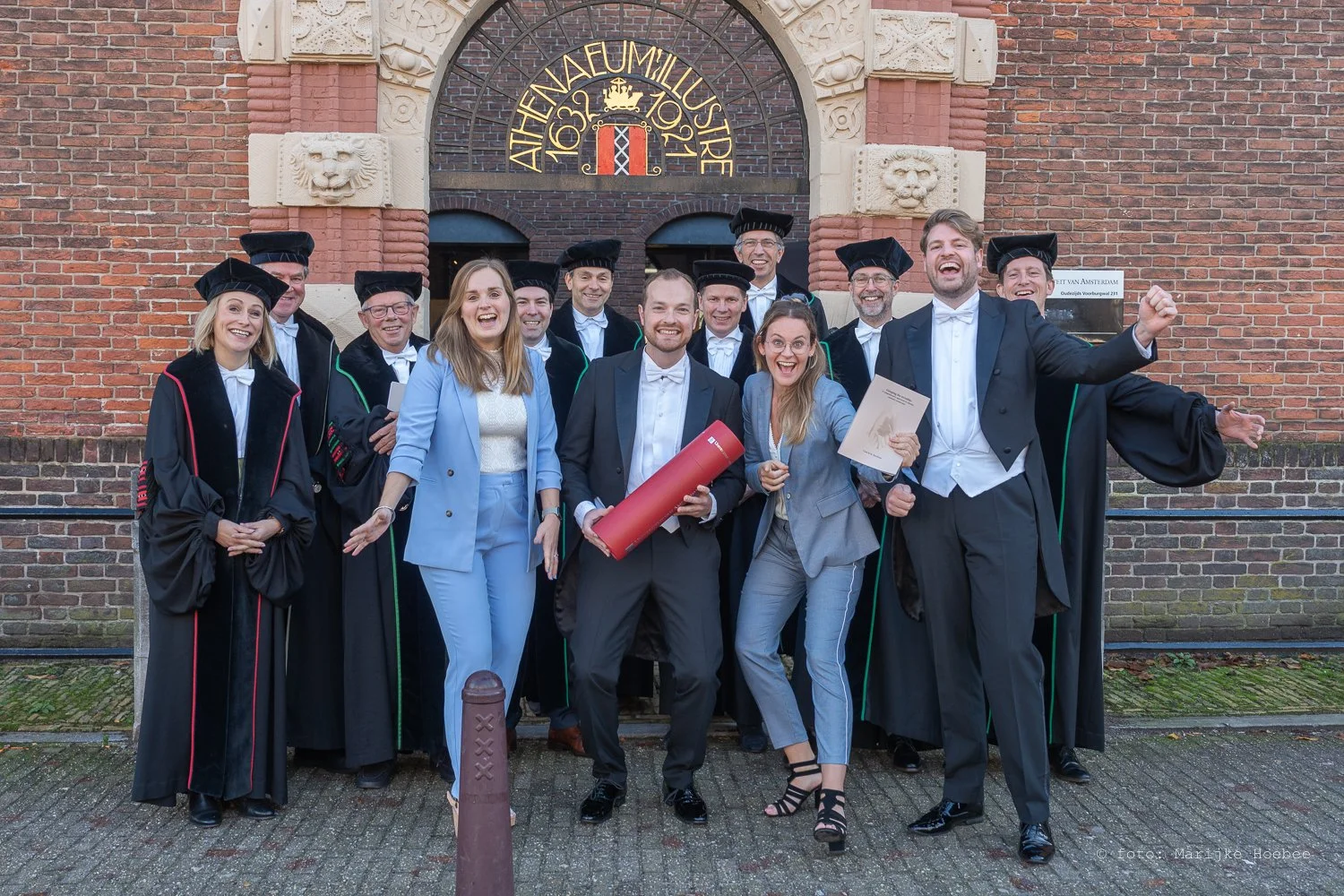 Groep afgestudeerden in academische robes en hoeden lachen en vieren voor een historische gevel met het jaar 1632. Ze houden diploma's en een diplomarol, symbool van hun afstuderen.