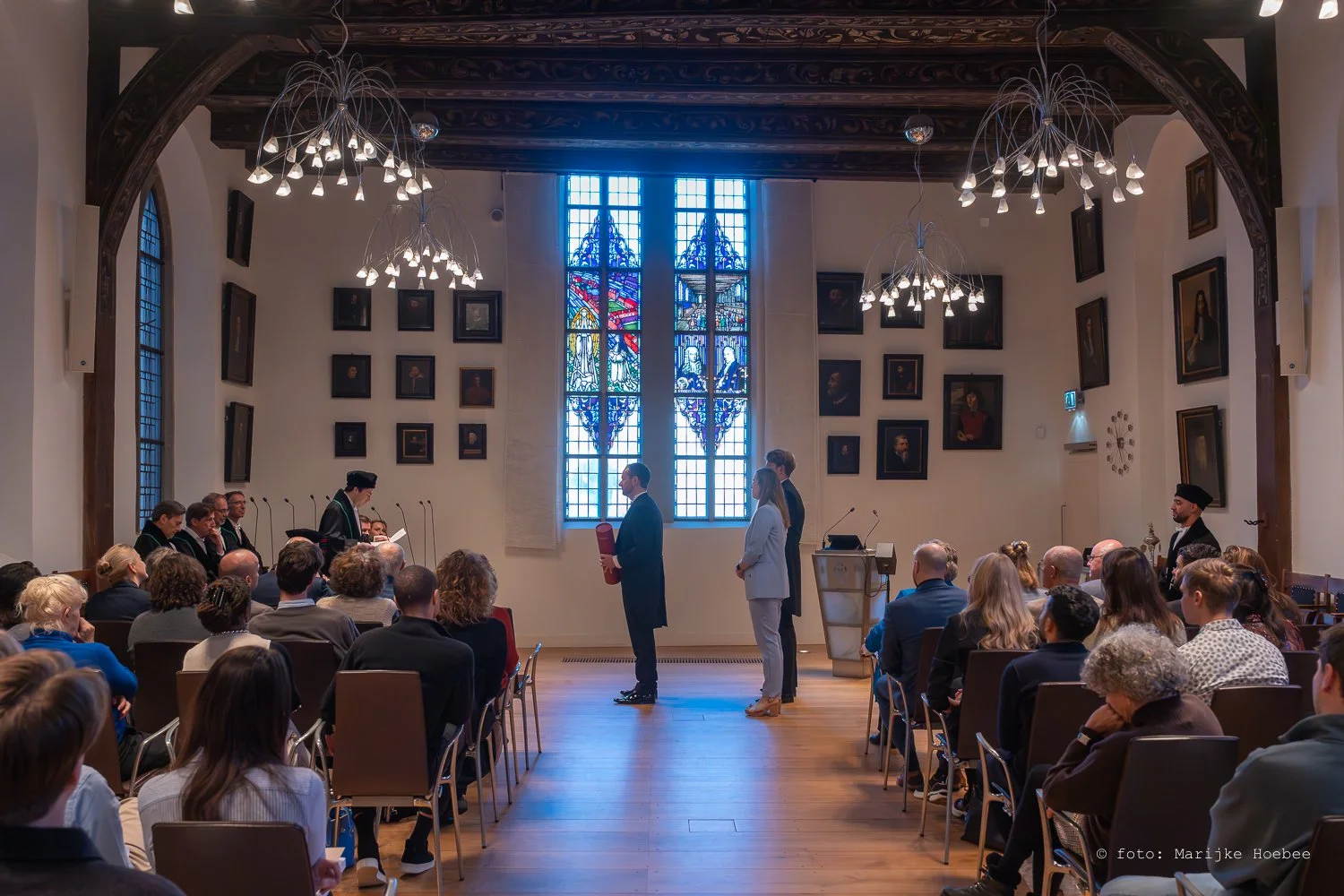Grote groep mensen in een grote, lichte zaal met hoog plafond en grote vitrale ramen, waarbij een ceremonie plaatsvindt met een persoon die een papiertje voorleest. Mensen zitten en kijken naar het toneel, met enkele personen die vooraan staan en een