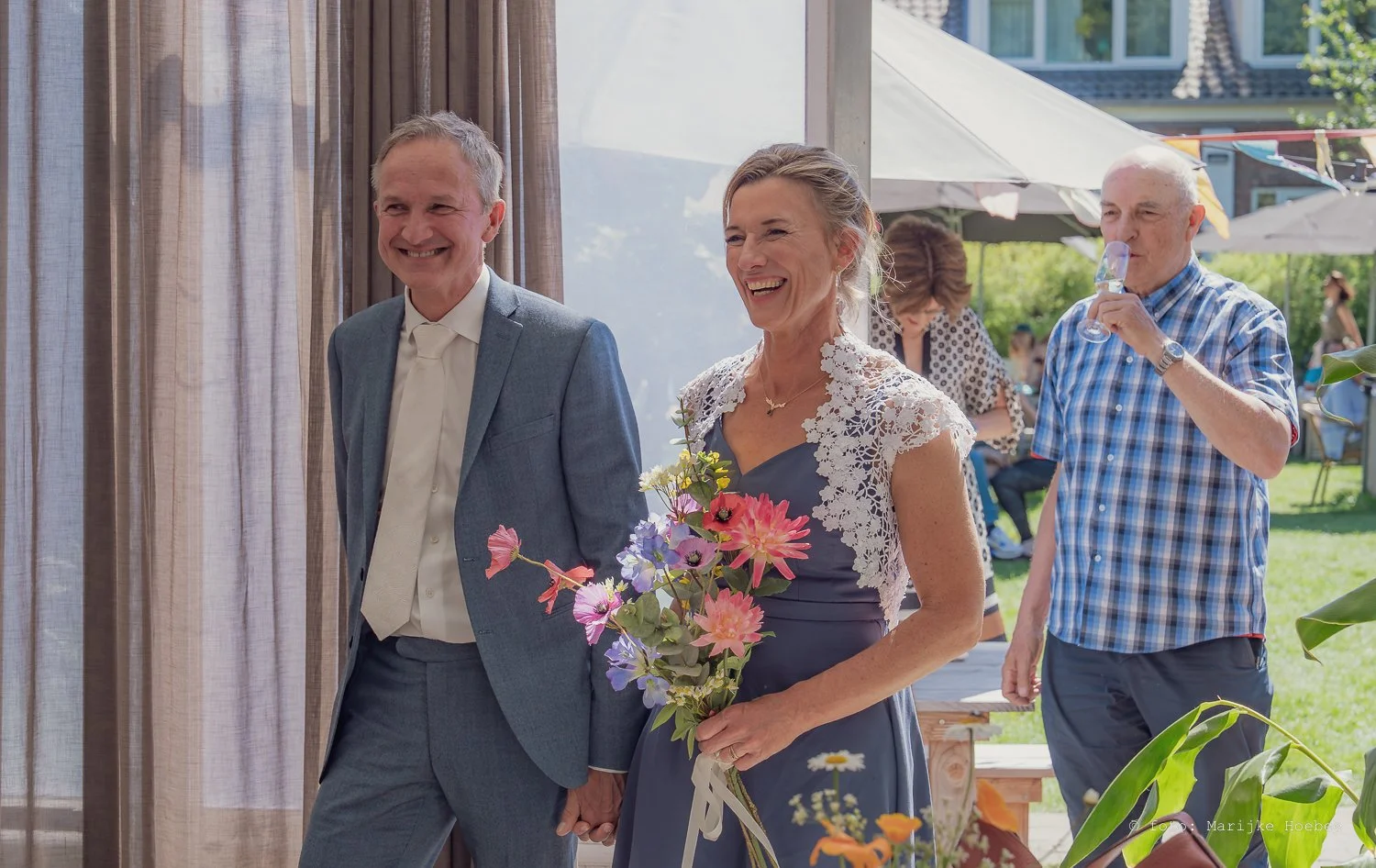 Een bruid en bruidegom lopen samen door een open tuin tijdens een buitenbruiloft, omringd door gasten en zonlicht.