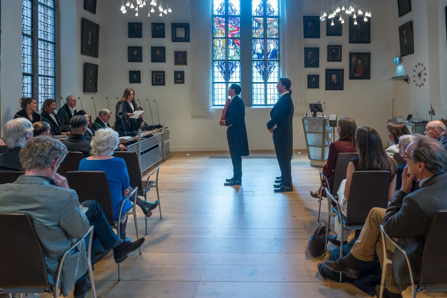 Een rechtszaak of ceremonie vindt plaats in een kamer met hoge ramen en een groot kunstwerk op de muur. Drie mannen staan voor de rechters, die rechts van het midden zitten. Mensen zitten in de zaal, enkele nemen notities.