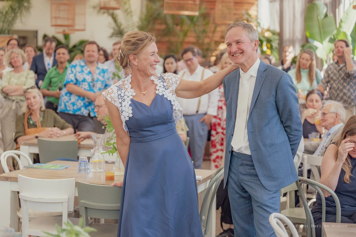 Een ceremonie of feest met een vrouw in een blauwe jurk met witte kant aan de schouders die lachend met een man in een lichtblauw pak met witte stropdas communiceert, omringd door aanwezigen in een groene, casual en formele outfit in een binnenruimte