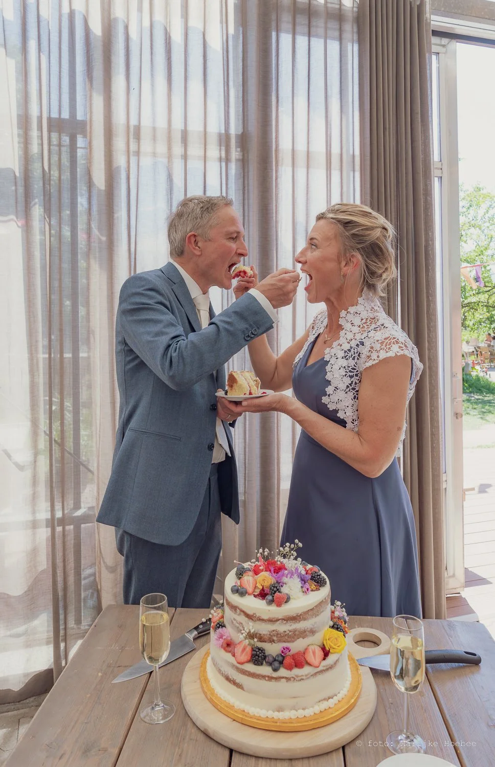 Een bruidspaar deelt een stuk taart tijdens hun huwelijk. Ze staan naast een tafel met een twee-laagse cake en twee glazen champagne.