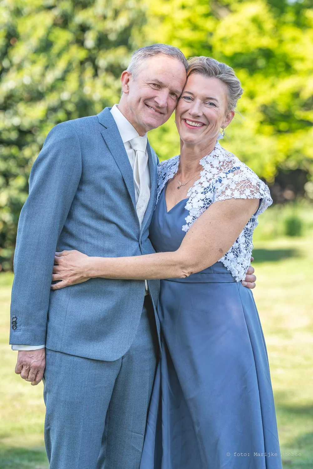 Een gelukkige oudere man en vrouw die elkaar omarmen, beiden in formele kleding, buiten op een zonnige dag met groene bladeren op de achtergrond.