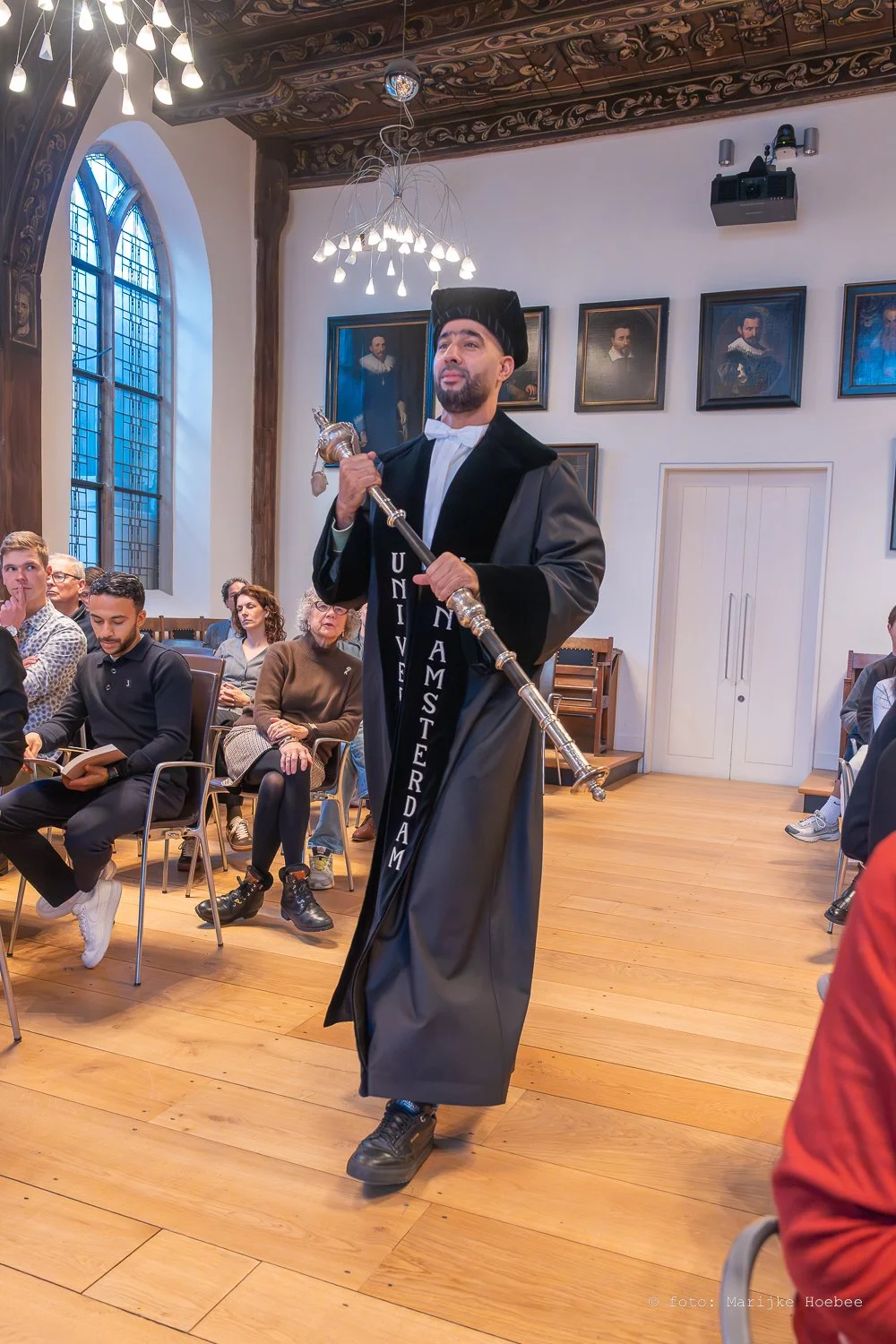 Man in academisch gewaad en baret houdt een staf in de hand, met een lint dat 'Universiteit Amsterdam' leest. Hij staat in een historische kastanjebruine zaal met hoge, gebrandschilderde ramen en portretten aan de muur, terwijl een publiek op rij zit