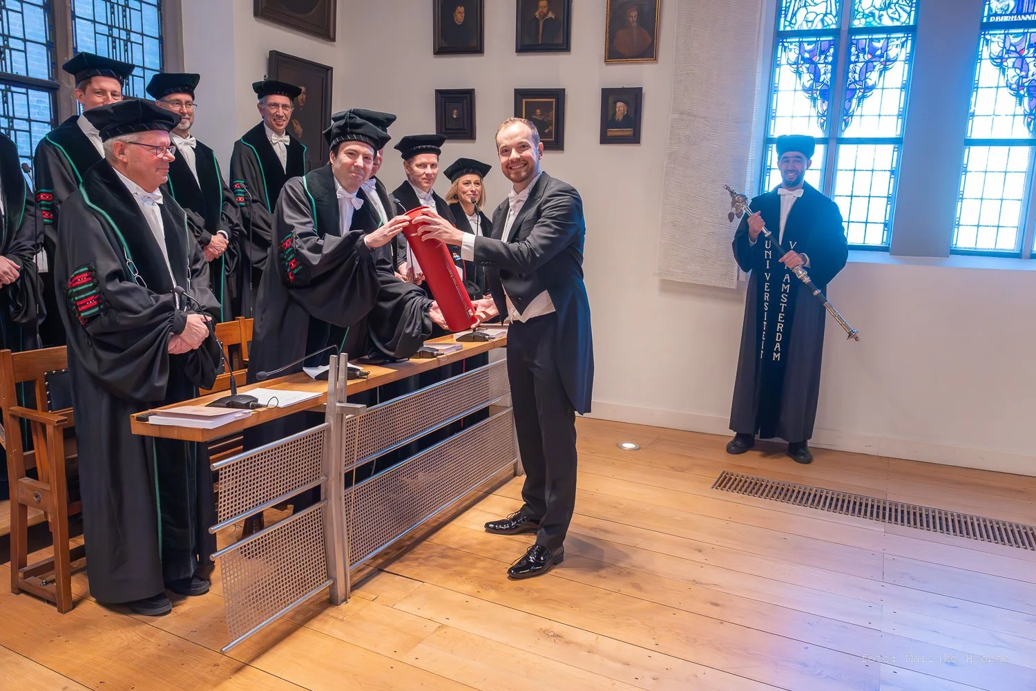 Een diploma-uitreikingsceremonie aan de Universiteit van Amsterdam, met meerdere afgestudeerden in academische gown en hooivork, en een man die het diploma ontvangt.