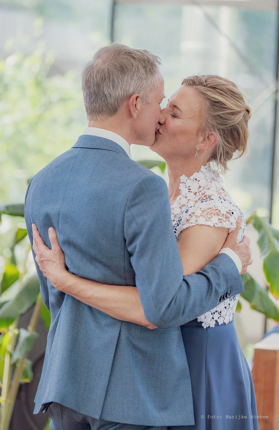 Een man en vrouw kussen elkaar liefdevol tijdens een speciale gelegenheid, waarschijnlijk een bruiloft, in een lichte ruimte met groene planten op de achtergrond.