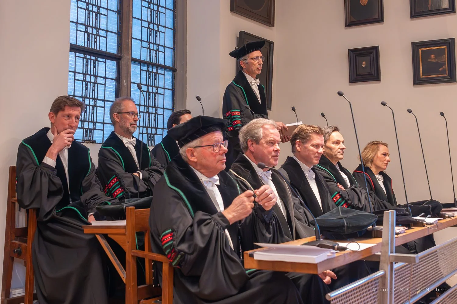 Juridische rechters en officieren in traditionele robes tijdens zitting in een historische kamer met grote glas-in-lood ramen.