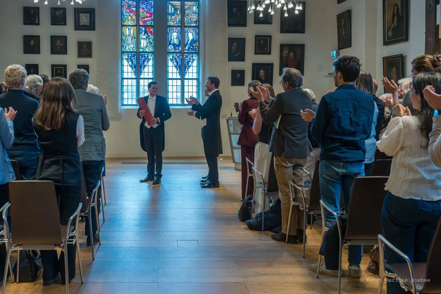 Een groep mensen in formele kleding staan en applaudisseren in een grote zaal met glas-in-loodramen en portretten aan de muur.