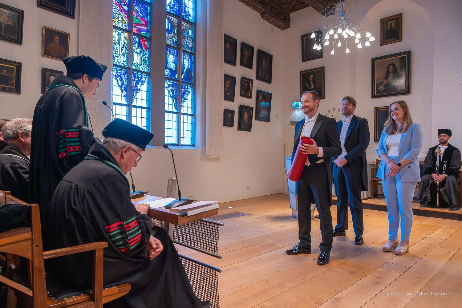Een formele prijsuitreiking in een kerk met een groot gekleurd glas-in-loodraam. Een man in pak ontvangt een certificaat of prijs uit een buis van een vrouw. Andere mensen in formele kleding staan achter hem, en enkele personen in traditionele kledin