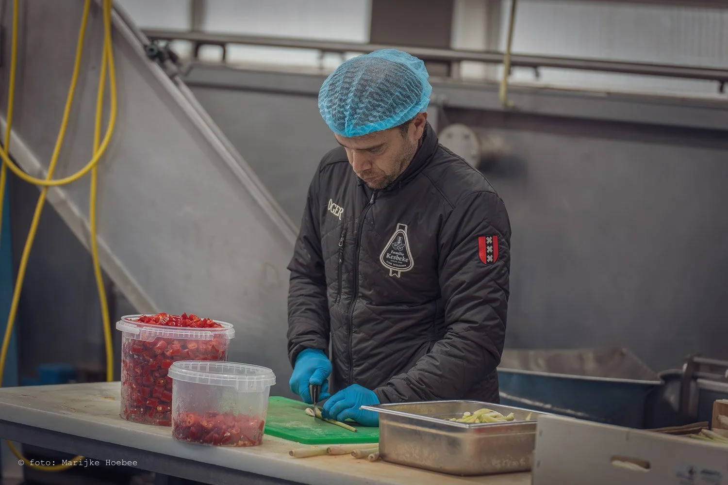 Man in een zwart jack met een blauwe haarnet en blauwe handschoenen snijdt groente op een groen snijplank in een fabriek of keuken.