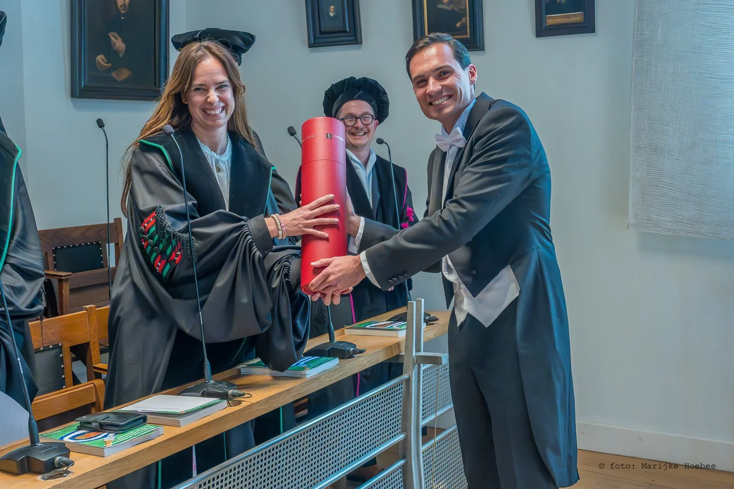 Een diploma-uitreiking met mensen in academische kledij, een vrouw wordt een diploma overhandigd door een man, op de achtergrond staan twee mannen in academische kleding.