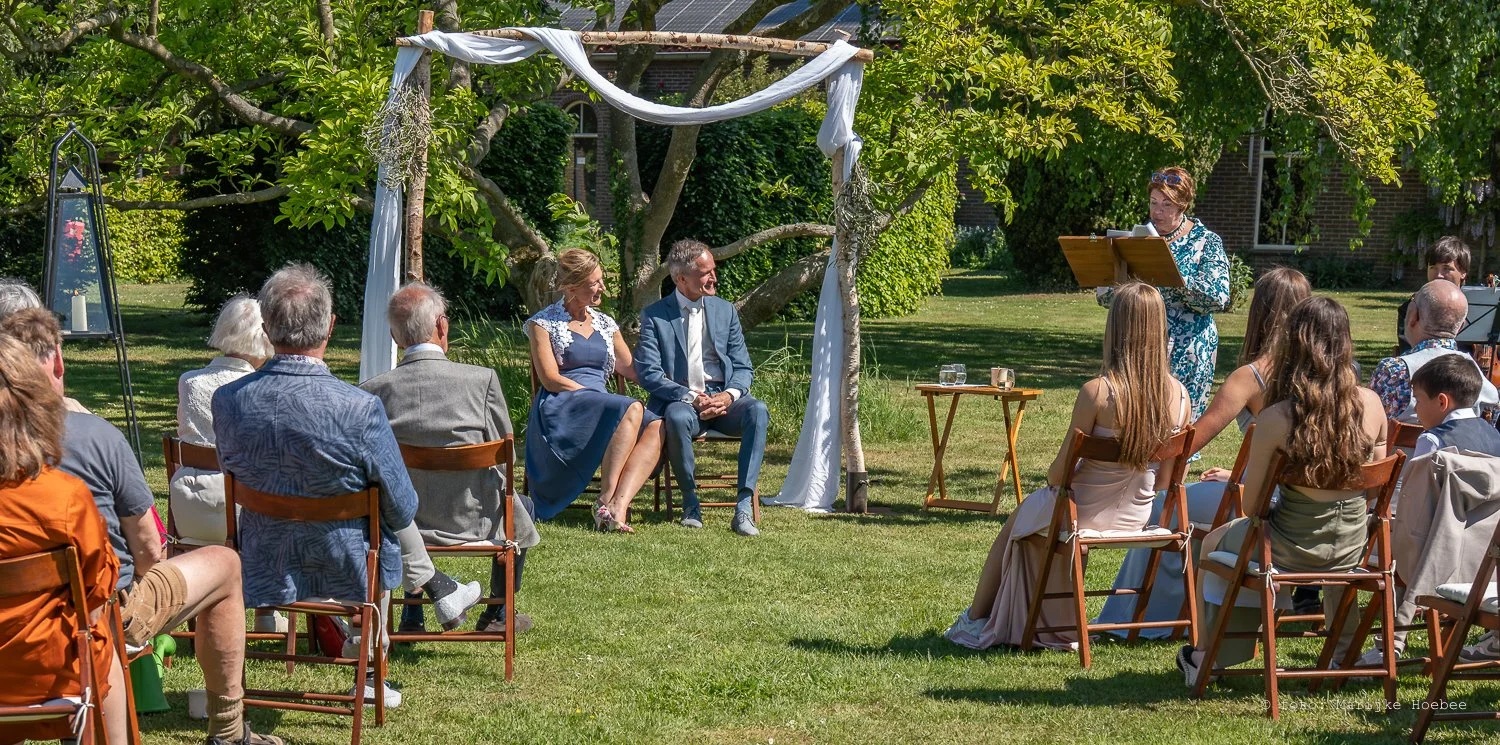 Een bruiloft buiten met een tent en een loper. Mensen zitten op stoelen en kijken naar een bruid en bruidegom die naast elkaar zitten, terwijl een vrouw voorleest of spreekt.