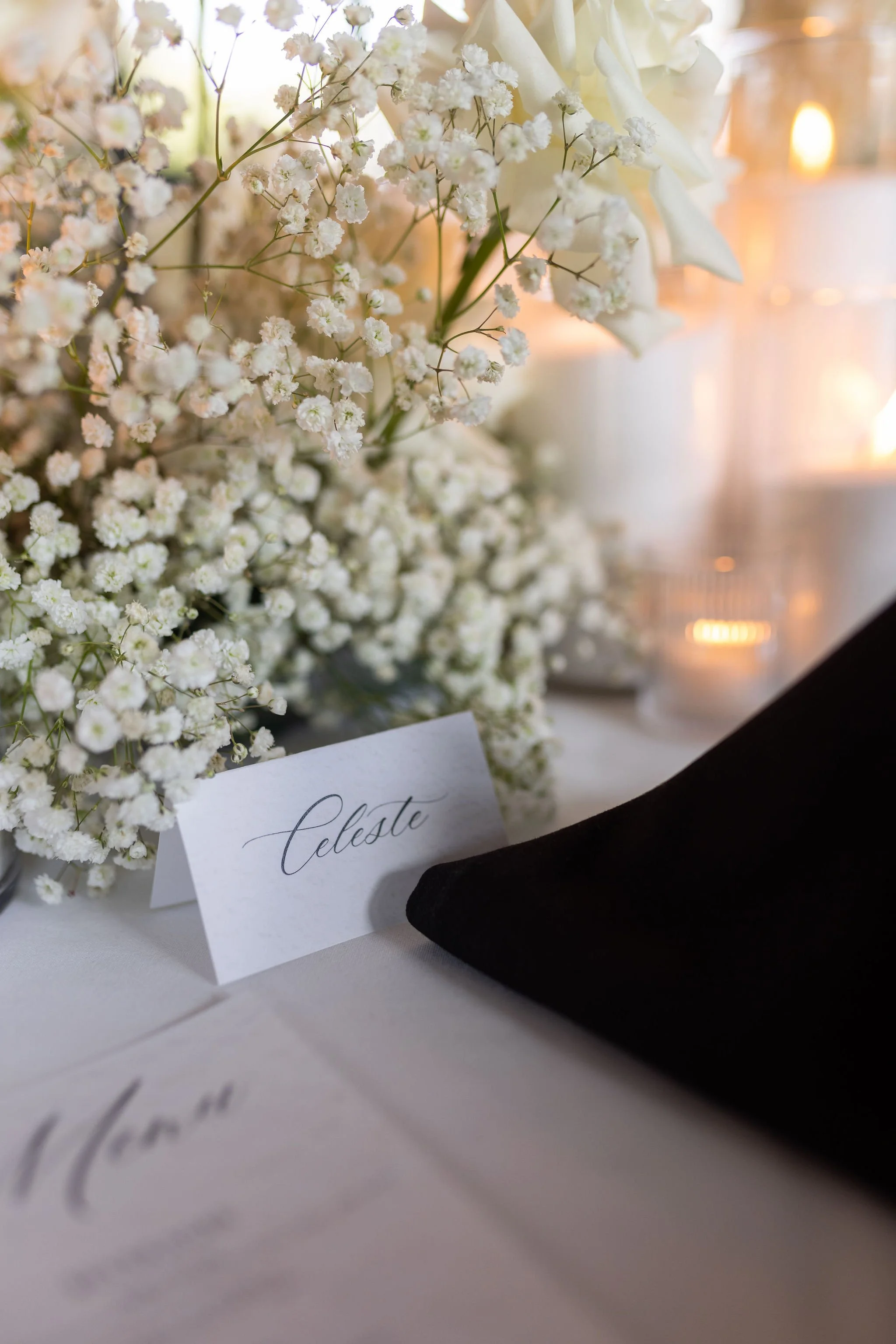 A close-up of a white floral arrangement with small white flowers and some larger leaves, placed on a table decorated with a black fabric. A white place card with elegant cursive writing that says 'Celeste' is positioned in front of the flowers. In the background, soft warm lighting creates a gentle glow, possibly indicating a wedding or elegant event setting.
