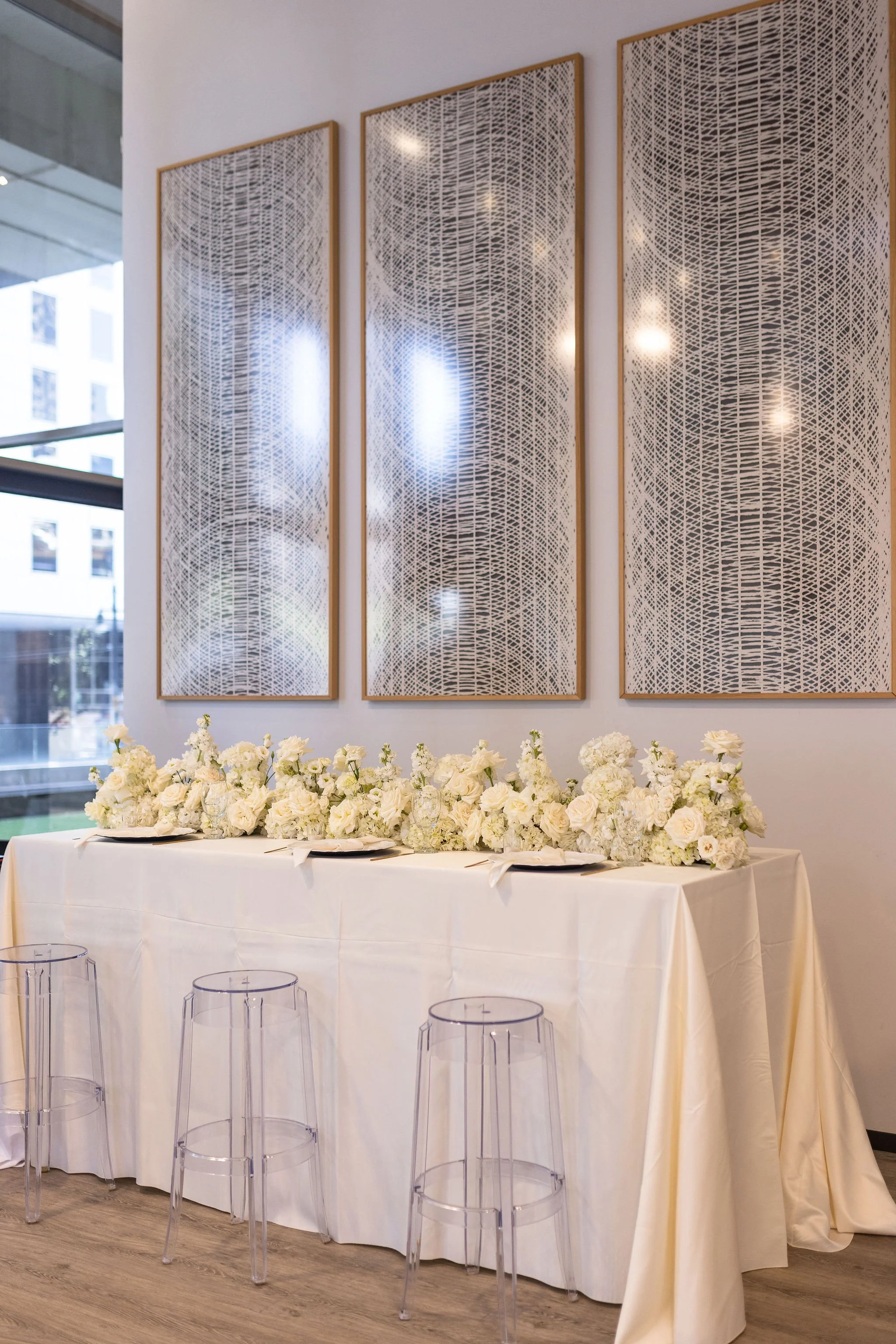 A long table covered with a white tablecloth, decorated with a large arrangement of white flowers, set for an event. Behind the table are transparent modern chairs, and in the background, three large abstract black-and-white framed art pieces hang on a white wall. Windows show neighboring buildings outside.