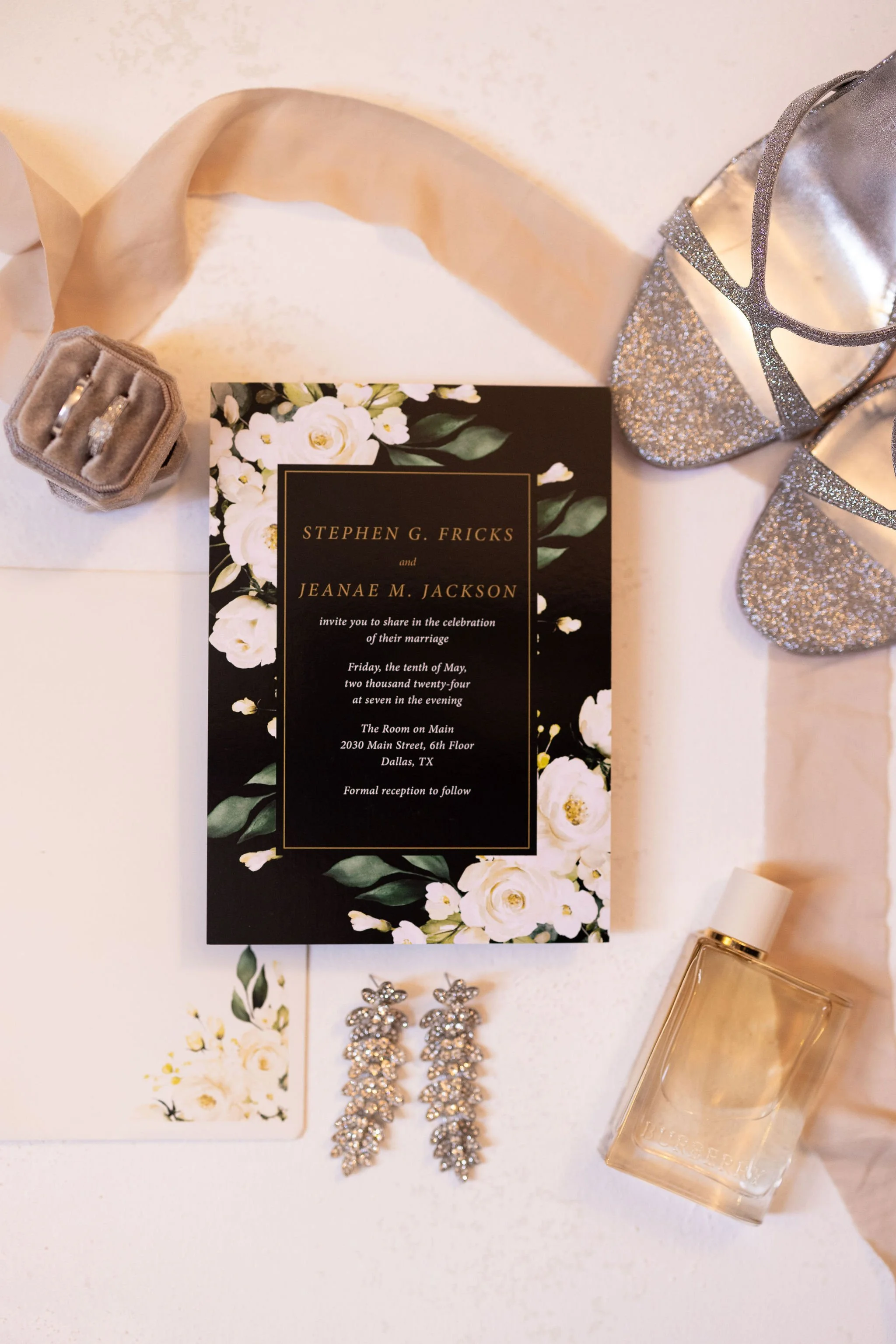 Wedding invitation surrounded by jewelry, perfume, and shoes on a white surface.