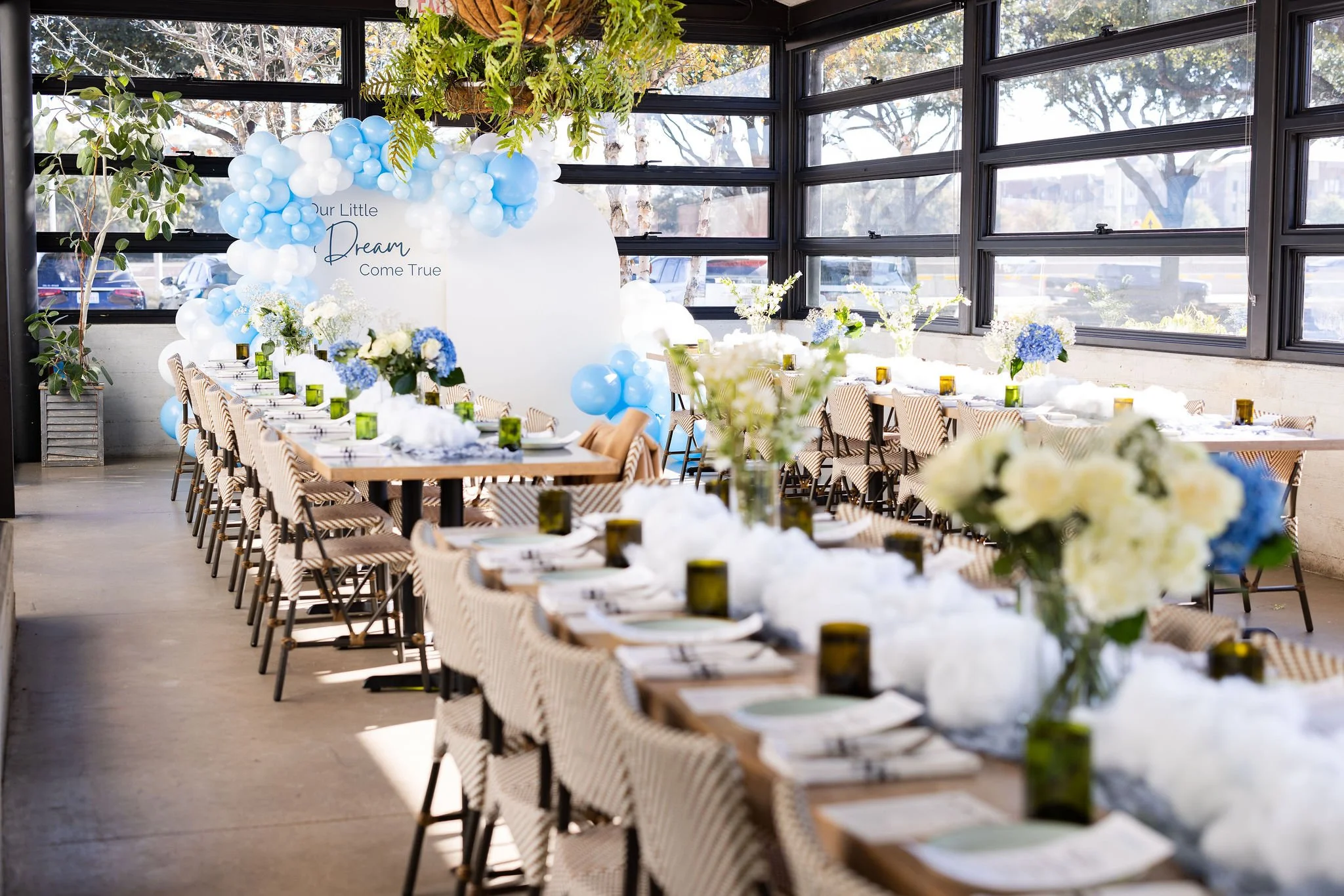 A decorated indoor event space with a long table set with plates, cups, and floral centerpieces, surrounded by chairs with striped cushions. The space has large windows with views of the outdoors and is decorated with white and blue balloons, flowers, and greenery, with a backdrop that reads "Our Little Dream Come True."