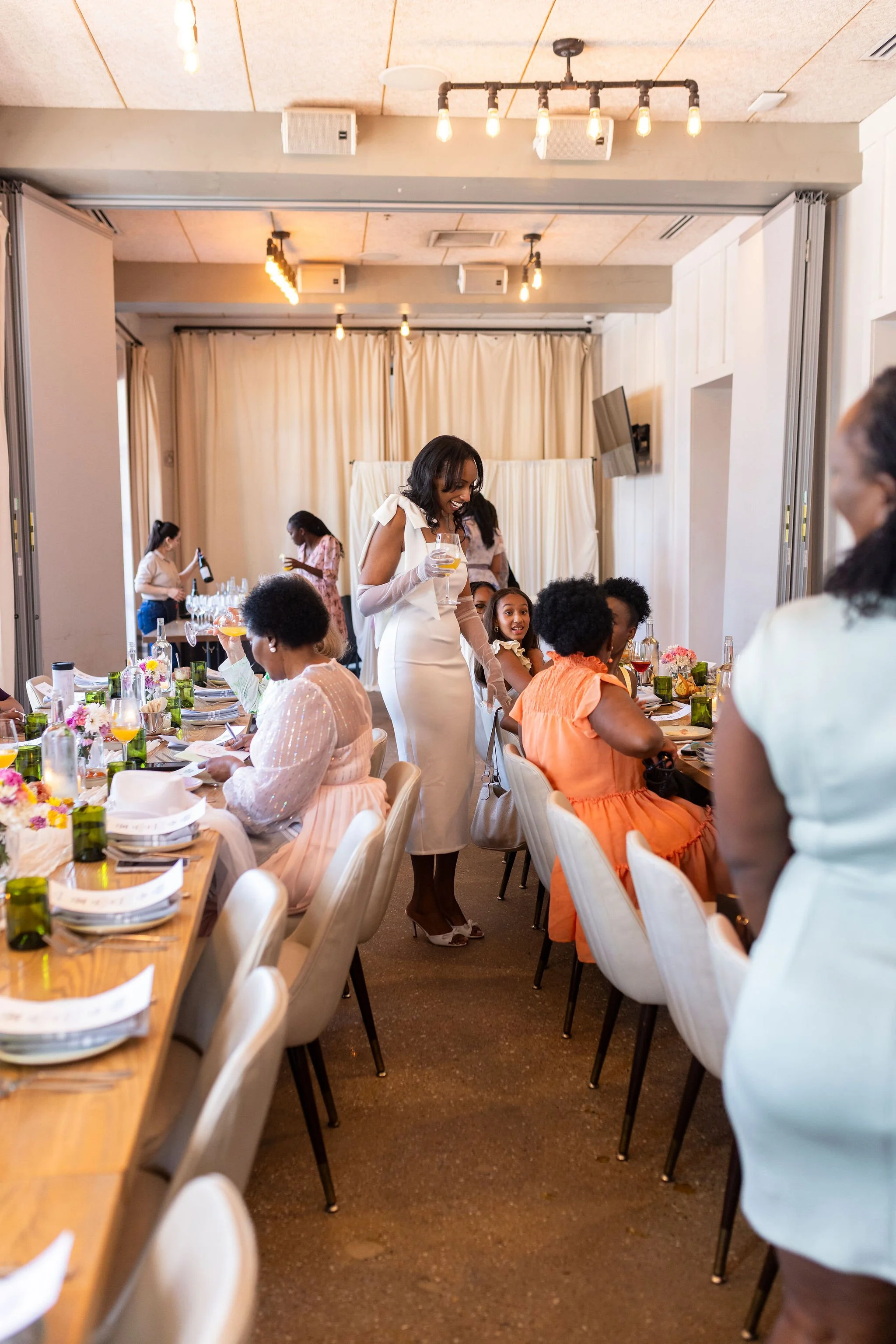 A woman in a white dress and gloves standing at a table, holding a glass of wine, smiling at a group of women seated at a decorated banquet table in a well-lit room with beige curtains and ceiling lights.