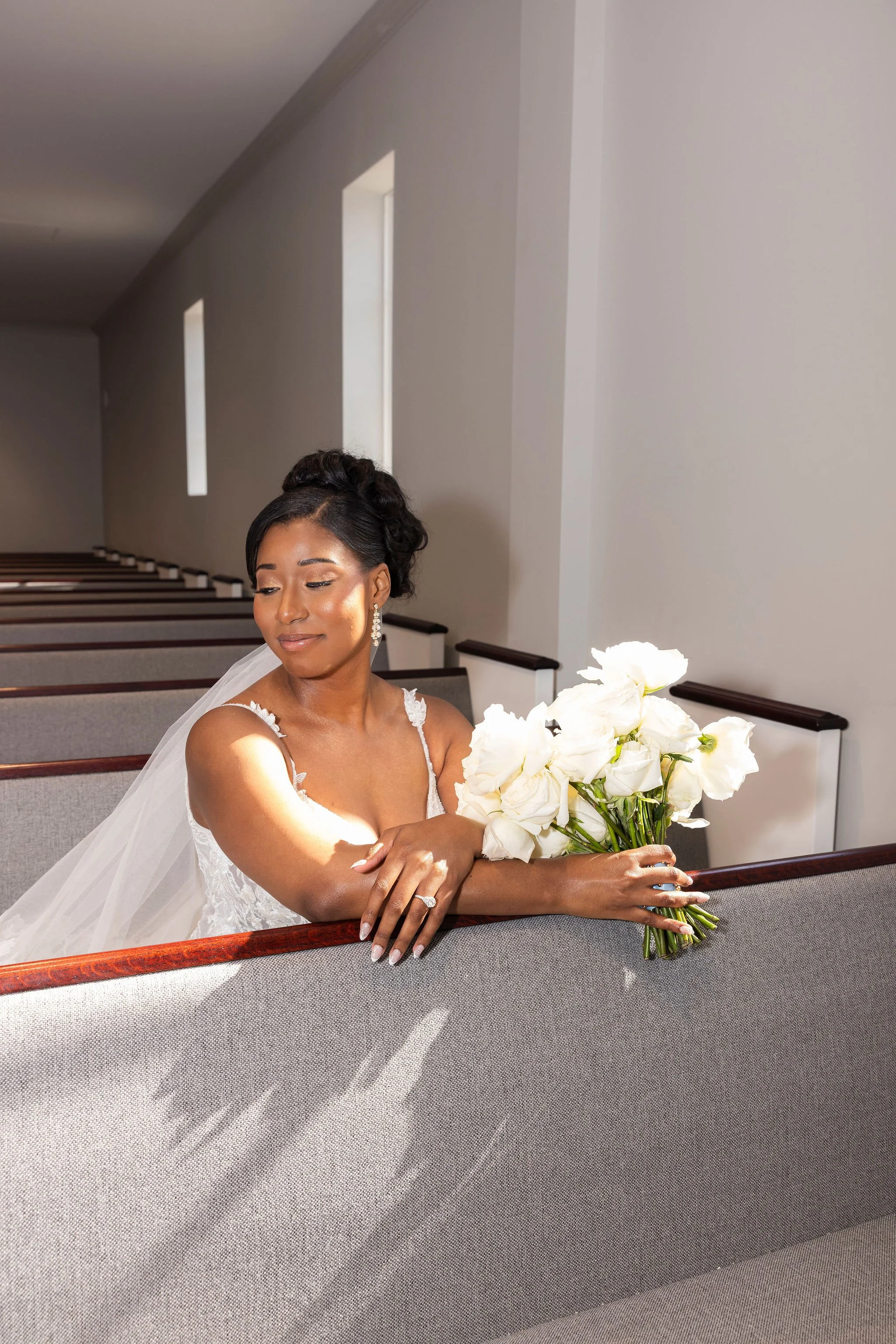 A bride in a white wedding dress with lace details and earrings, sitting in a church pew, holding a bouquet of white roses and greenery, with her eyes closed and a serene expression.
