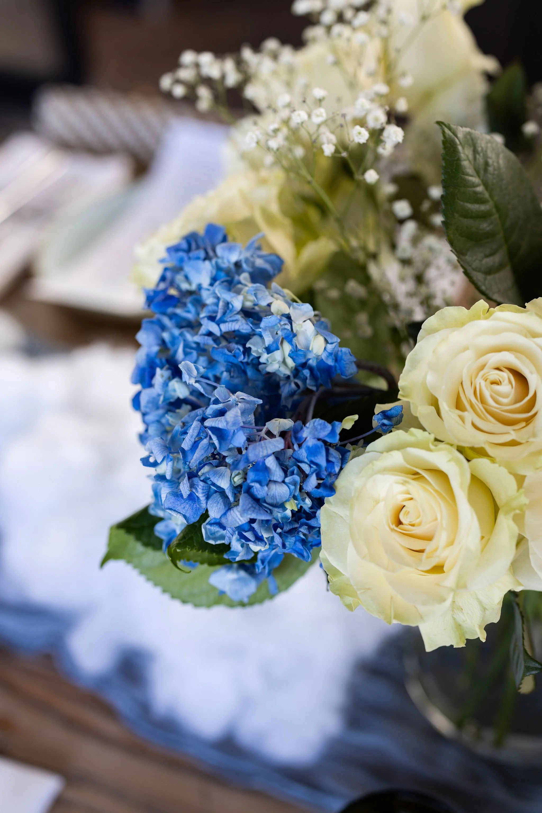 A close-up of a bouquet of flowers featuring yellow roses, blue hydrangeas, white baby's breath, and green leaves.
