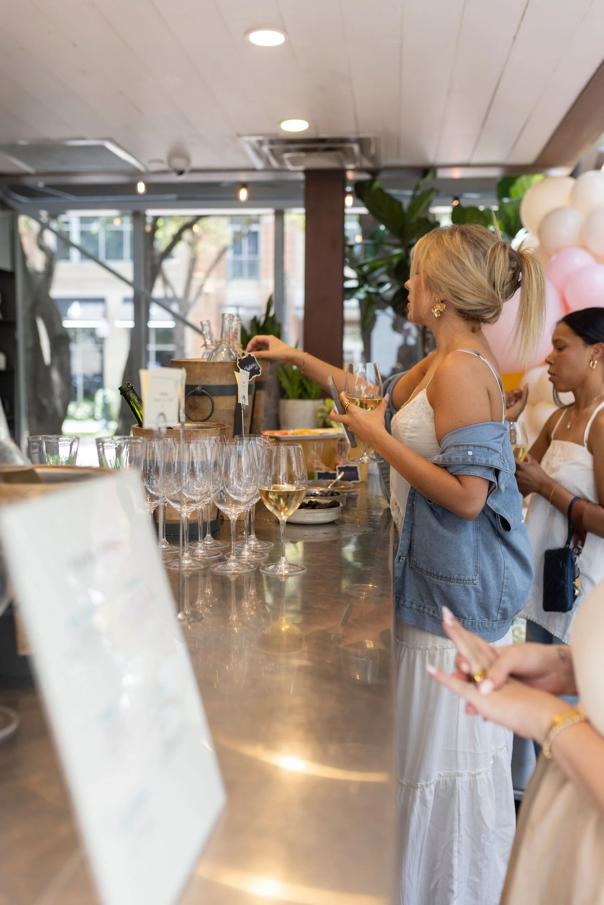 Women at a bar or cafe, serving and drinking wine, with glasses and decor around, in a bright indoor setting