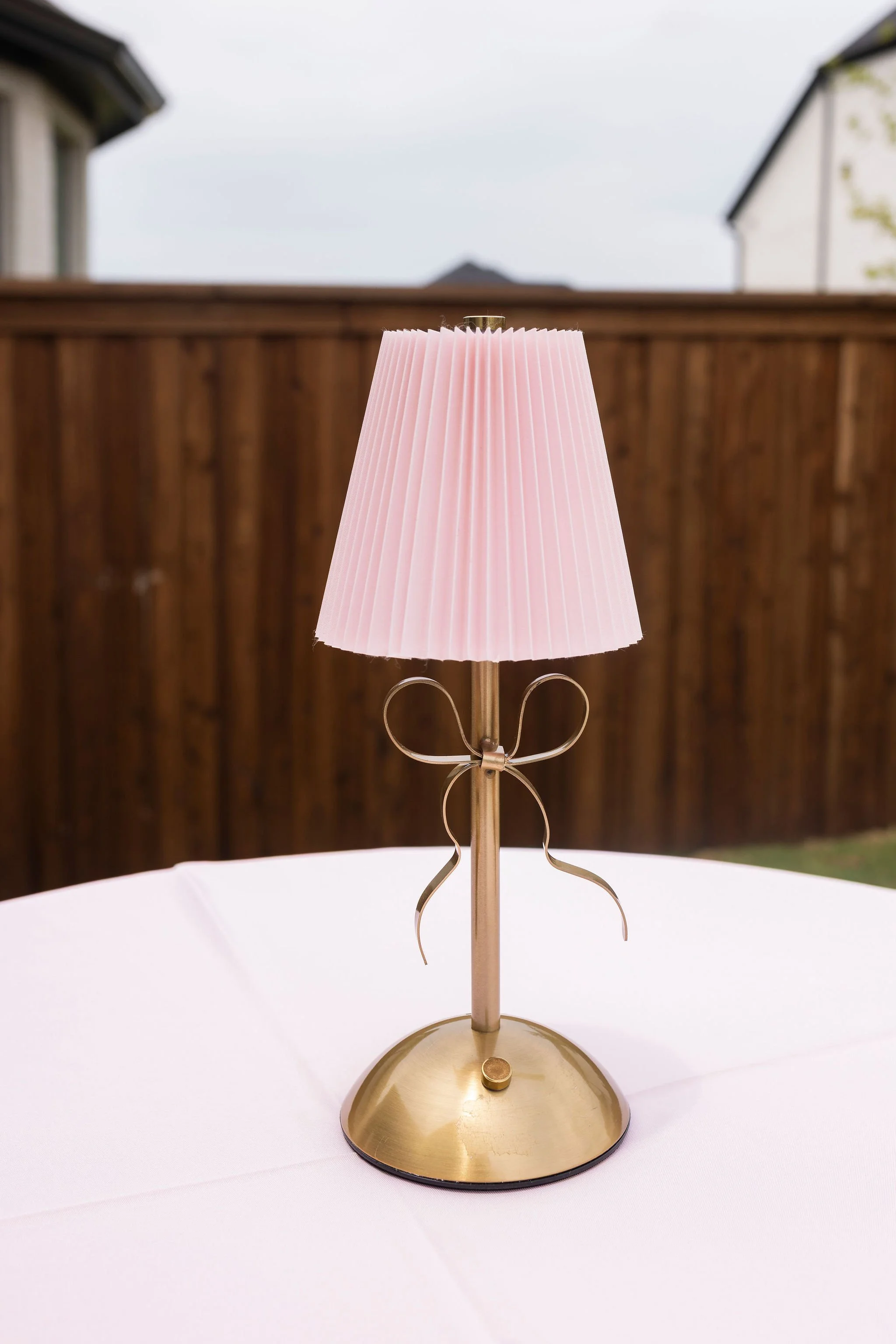 A brass table lamp with a pink pleated lampshade on a white tablecloth, outdoors with a wooden fence and houses in the background.