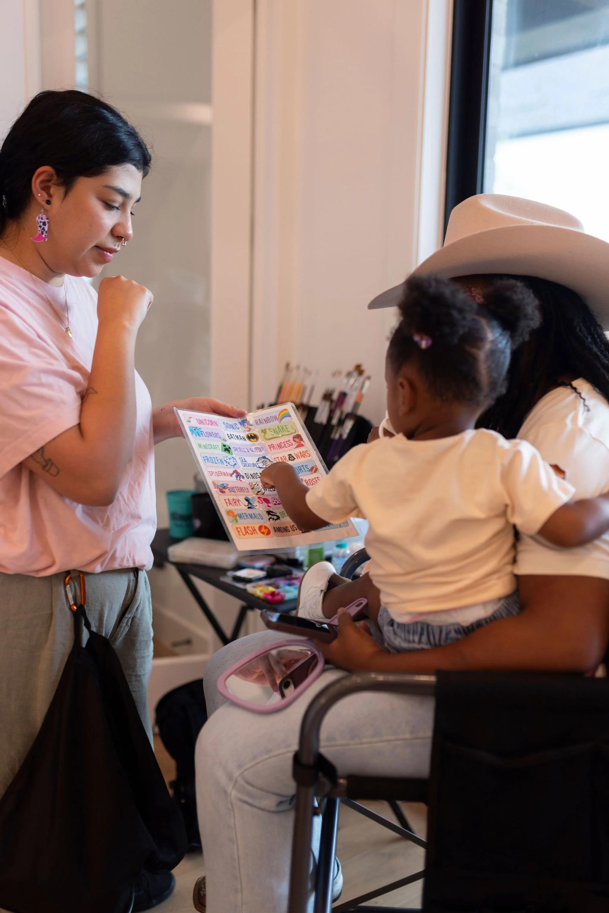 A woman with dark hair and earrings is showing a colorful sticker sheet to a young girl seated in a wheelchair. The girl wears a large wide-brimmed hat, a white shirt, and shorts, and is holding a tablet. They are indoors near a window, with a table 