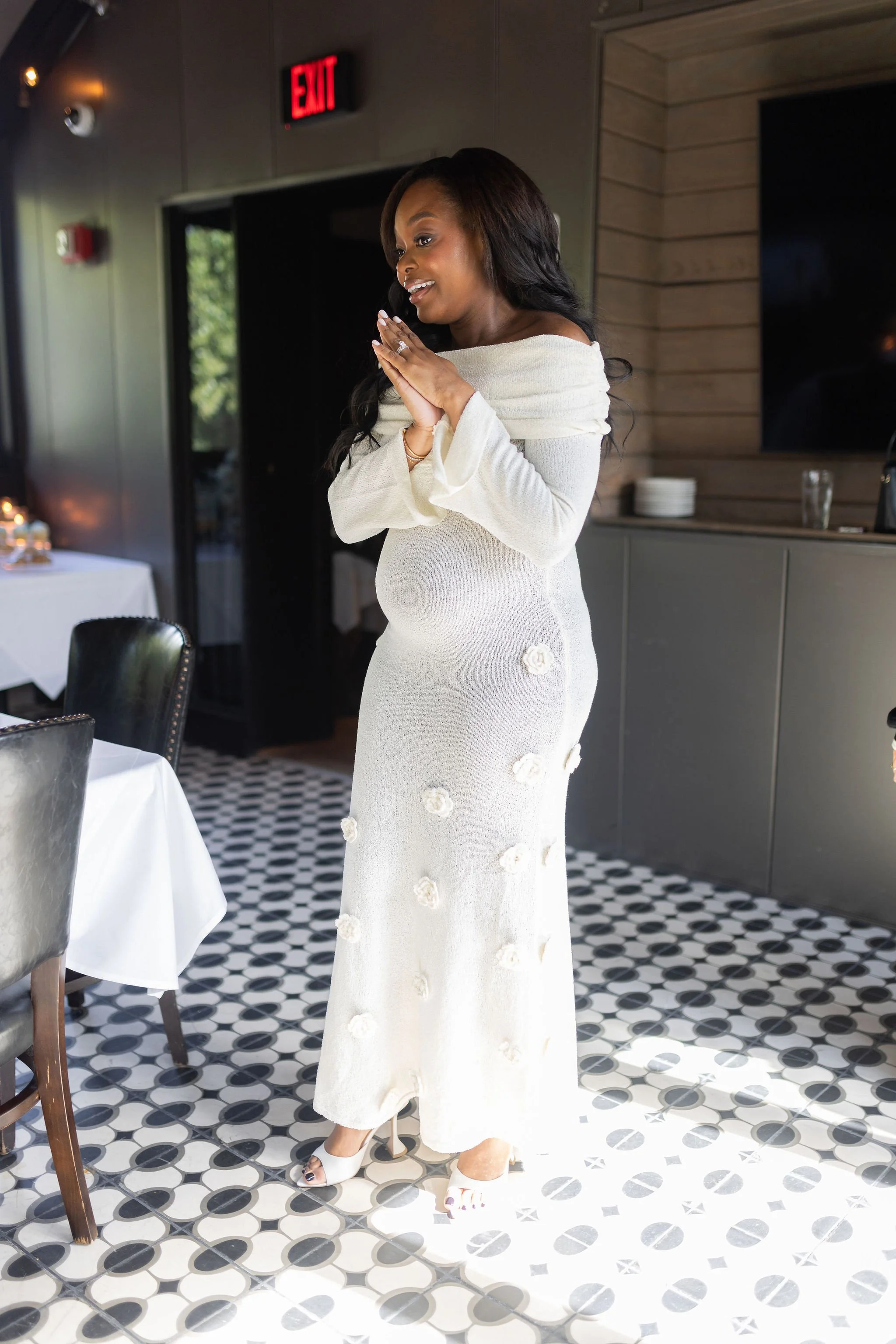 Woman in a white dress with flower details, standing inside a room with black and white patterned floor tiles, near a table with a white tablecloth, and a wall with wooden paneling and a television.