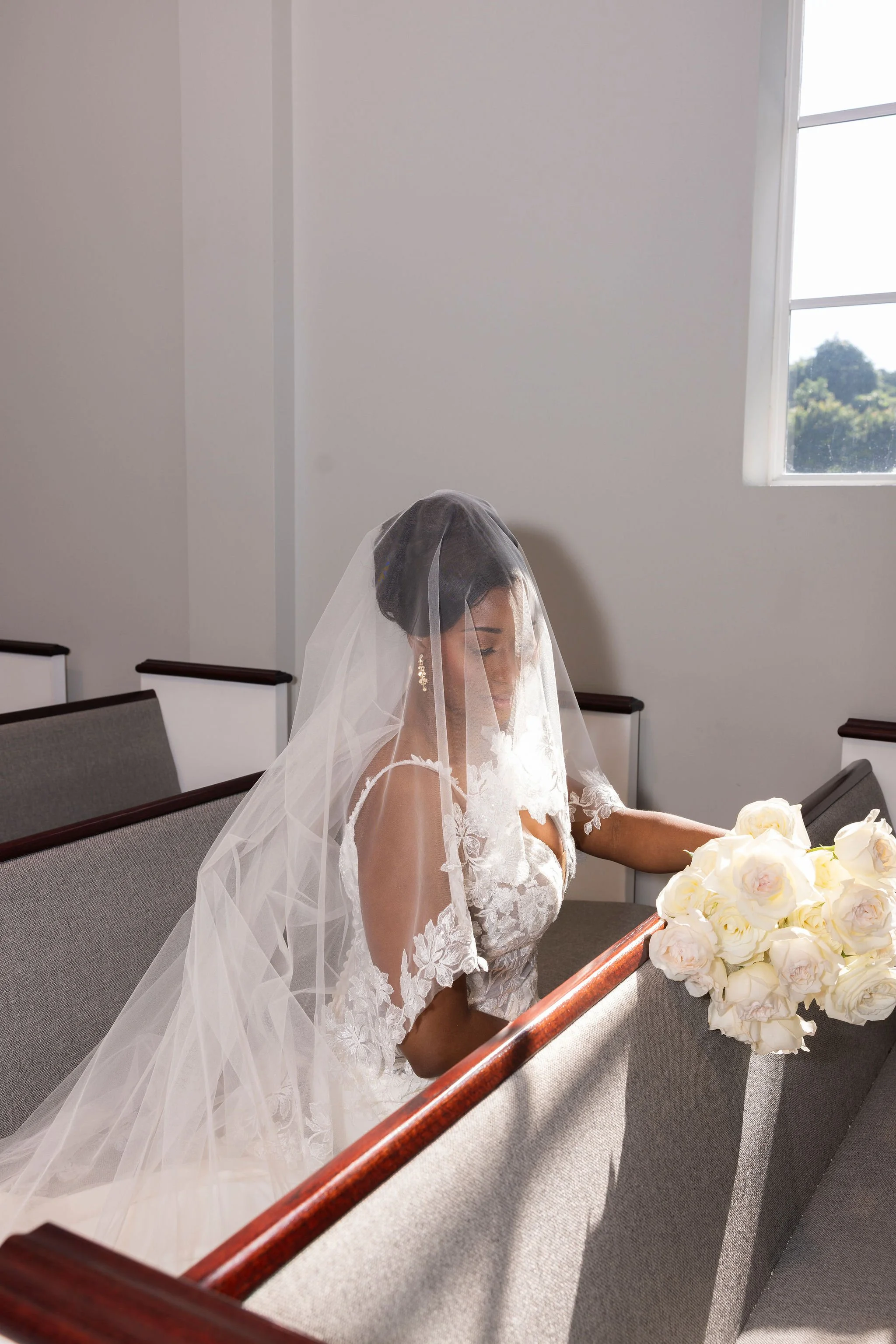 Photo of a bride in a white lace wedding dress with a veil, sitting in a church pew, holding a large bouquet of white roses, with natural light coming through a window.