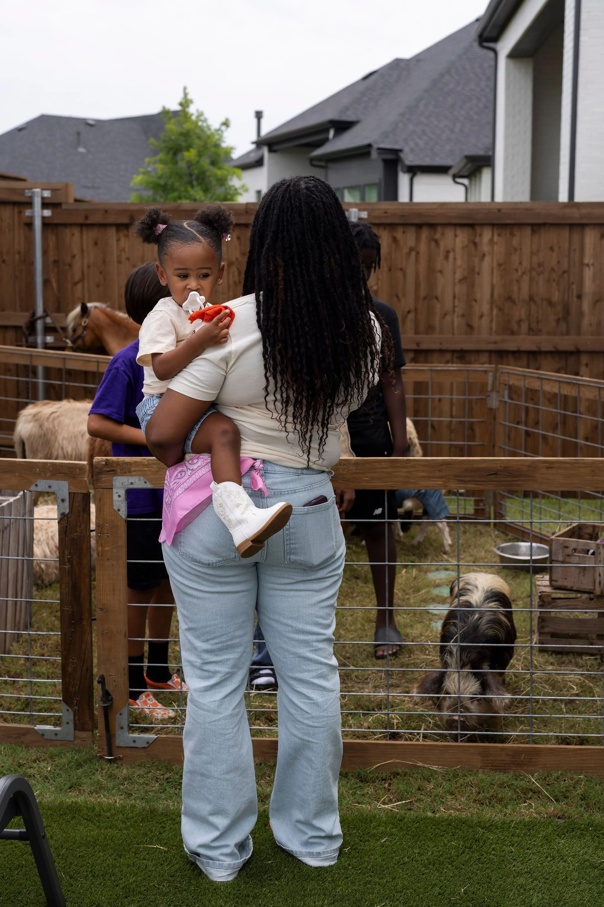 A woman holding a young girl near a wooden fence at an outdoor petting zoo. The girl has a pacifier, bunny ears in her hair, and is wearing a beige shirt and pink pants. There are goats and a dog in the enclosure with a farm-style setting, including 