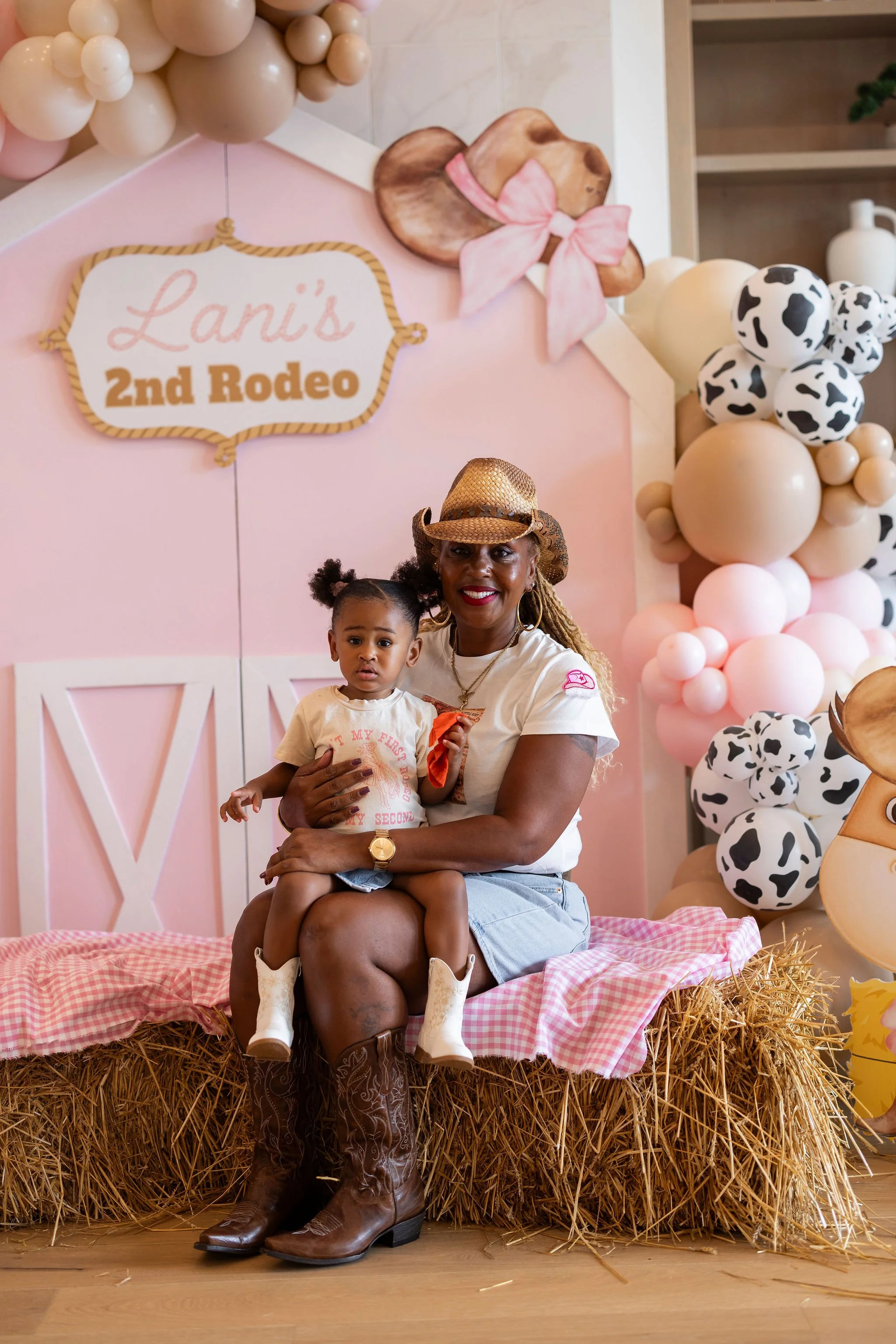 A woman and a young girl sit on a hay bale decorated with pink and white checkered fabric at a country-themed event. The woman is wearing a straw cowboy hat and cowboy boots, smiling at the camera. Behind them is a pink backdrop with a sign reading '