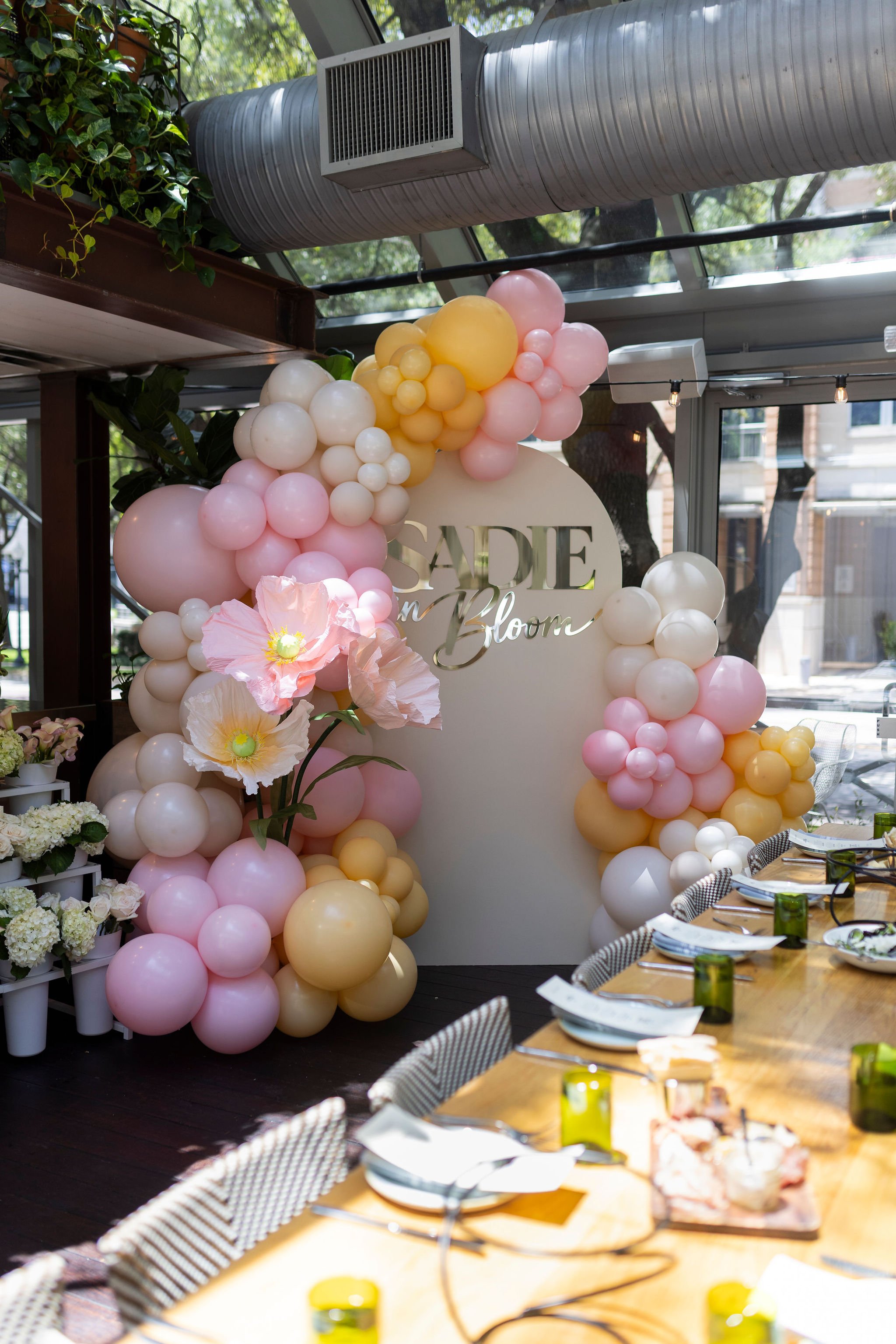 Decorated event space with pink, white, and yellow balloons arranged around a circular sign that reads 'Sade in Bloom.' The sign is partially visible behind flowers and balloons, with tables set for a meal with white plates, green glasses, and stripe