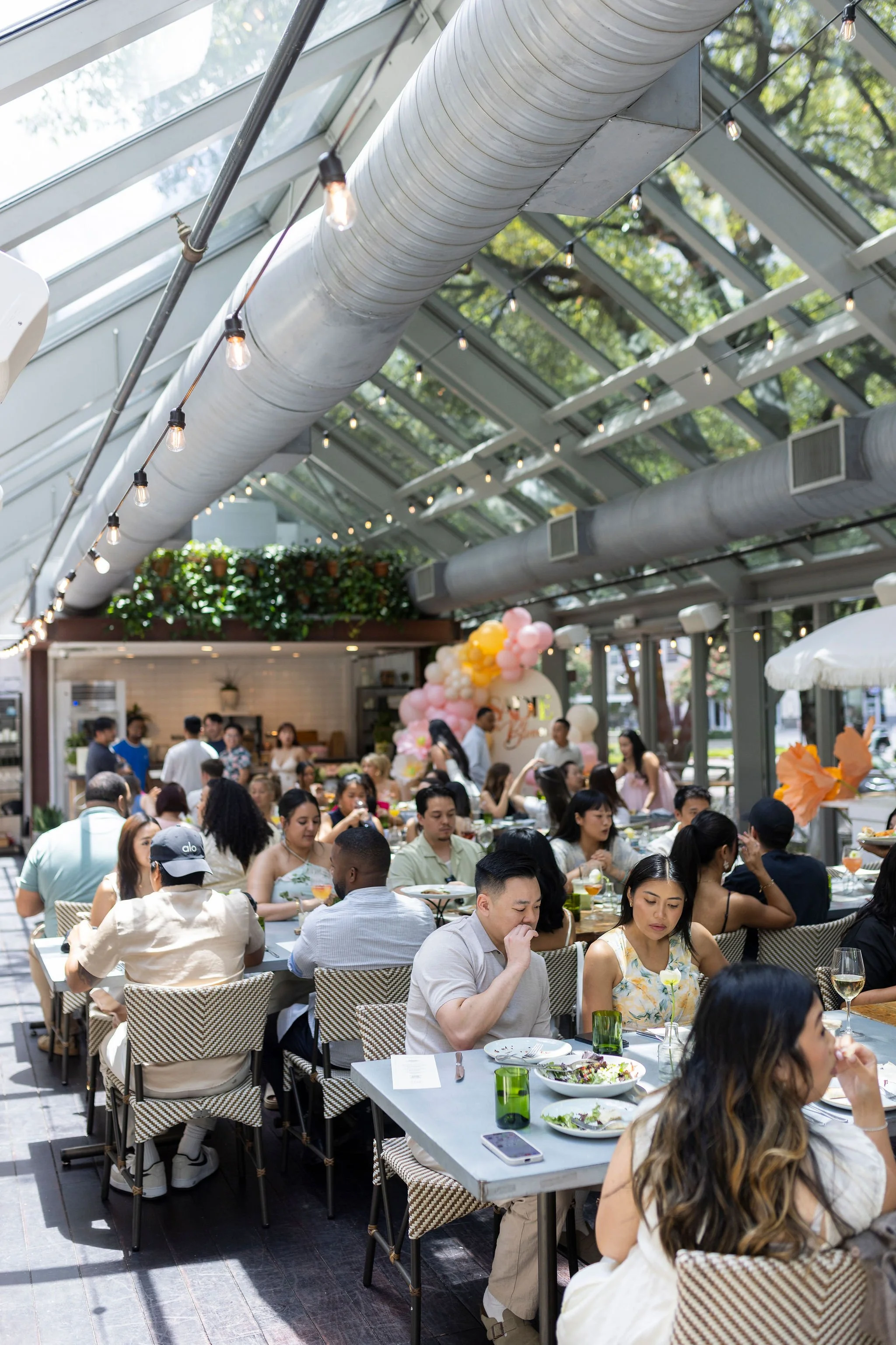 A crowded indoor restaurant with large glass windows and a glass ceiling, decorated with string lights and plants. Many people are seated at tables enjoying meals, with some engaging in conversation.