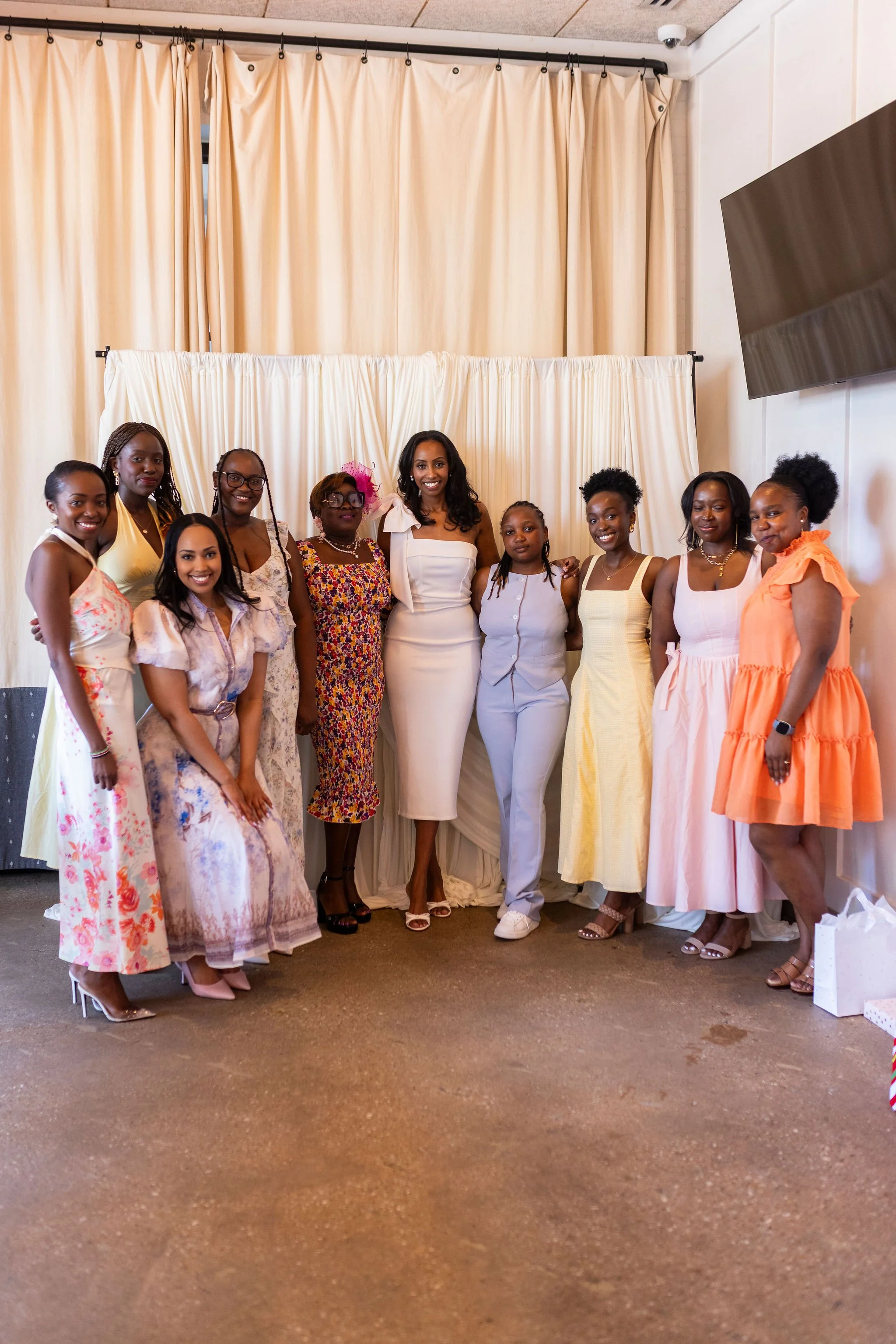 Group of women posing together in front of cream-colored curtains, dressed in colorful and elegant dresses for a celebration or event.