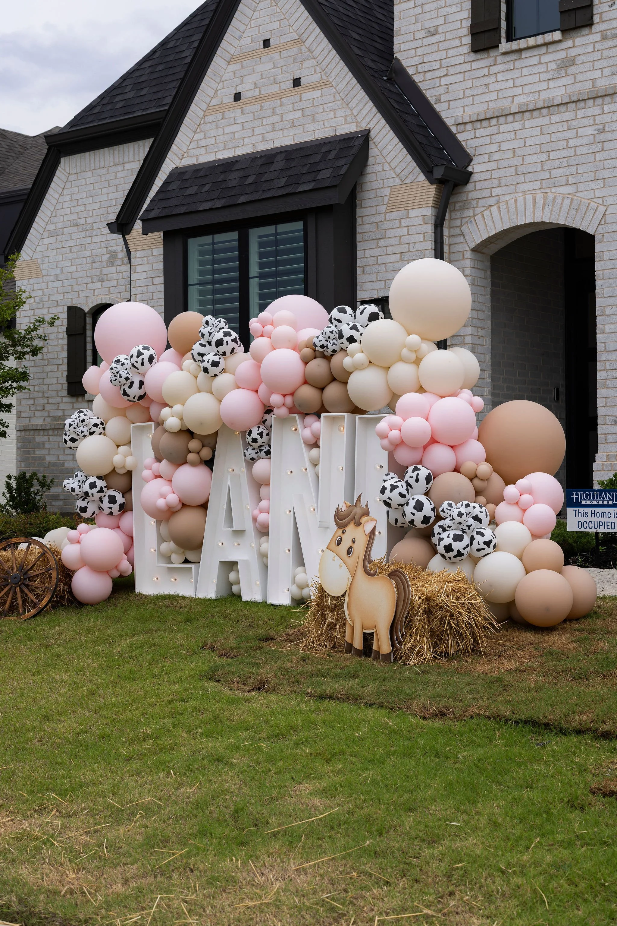 Decorative setup in front of a brick house featuring numerous pastel pink, white, tan, and cow print balloons clustered together. There are wooden letter signs and a cardboard cutout of a horse, with hay bales nearby, suggesting a farm or cowboy them