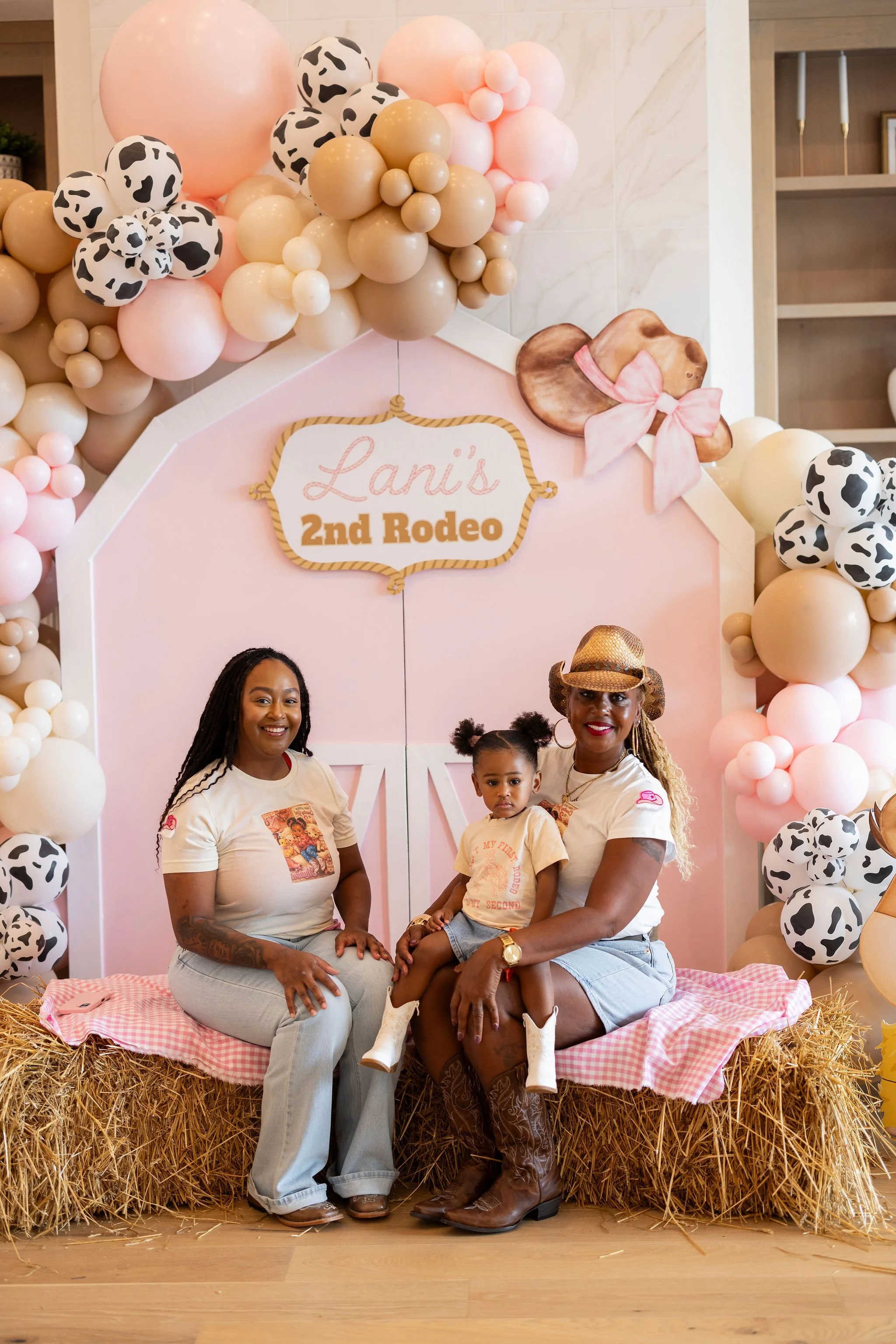 A family portrait with two women, a girl, and a boy sitting on a hay bale decorated with pink and white checkered cloth. Behind them is a pink backdrop with a sign that says "Lani's 2nd Rodeo" and a cowgirl hat hanging on a dog drawing with a pink bo