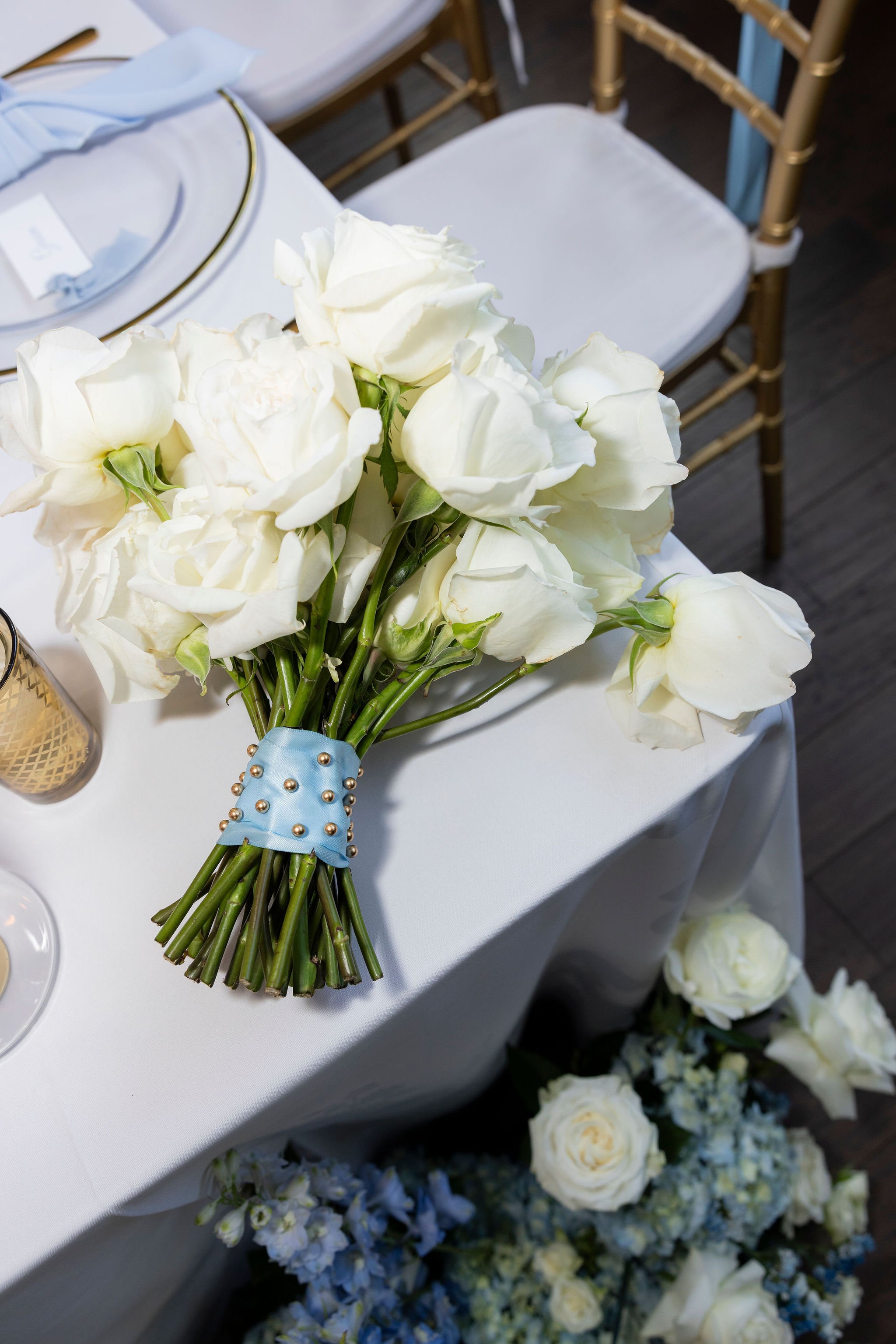 A bouquet of white roses with a blue ribbon and pearl accents resting on a table, with additional white and blue flowers beneath the table.