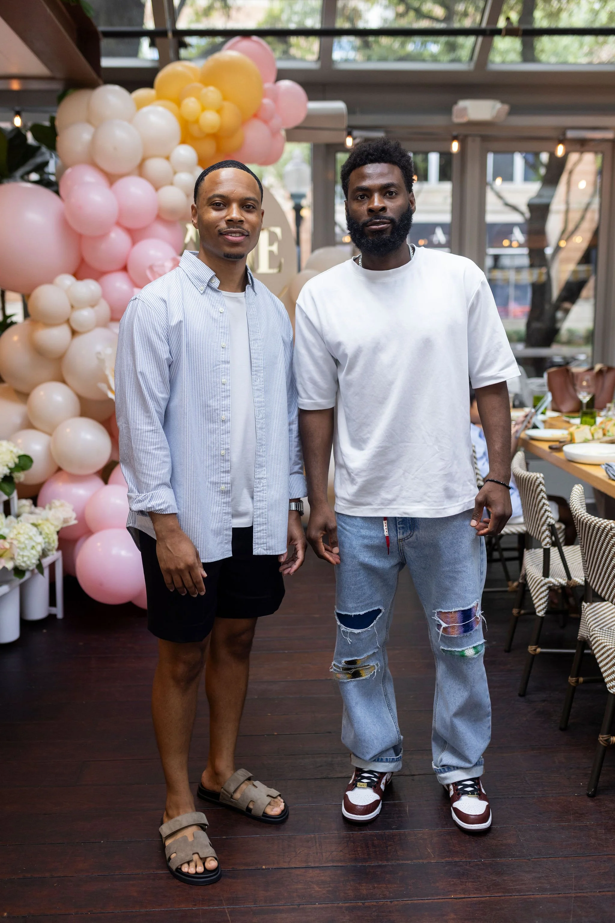 Two men standing indoors, one in a light blue striped shirt and black shorts, the other in a white t-shirt and ripped jeans, with a pink and white balloon decoration behind them.