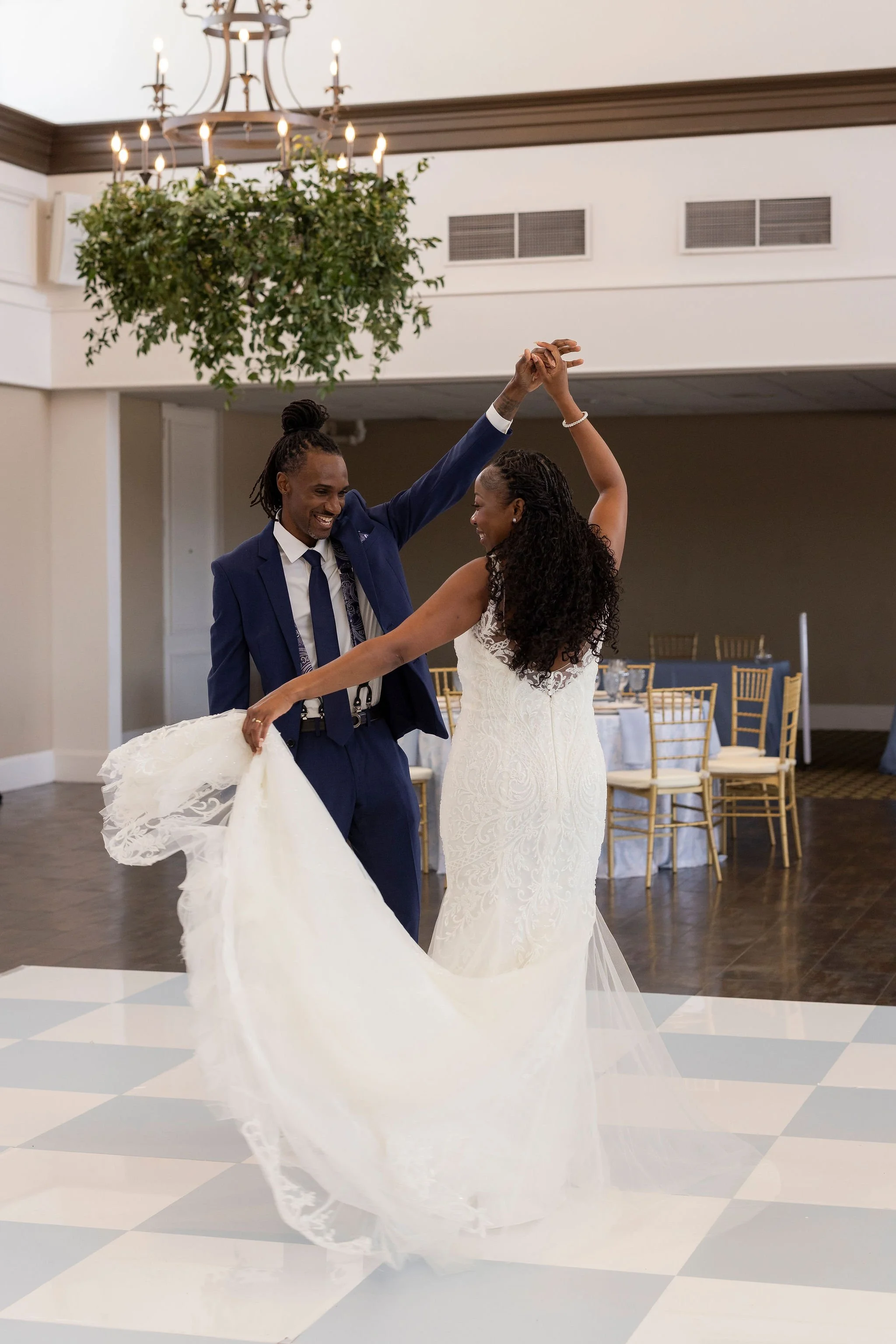 A bride and groom dancing at their wedding reception in a decorated hall, with the bride wearing a white lace gown and the groom in a blue suit.