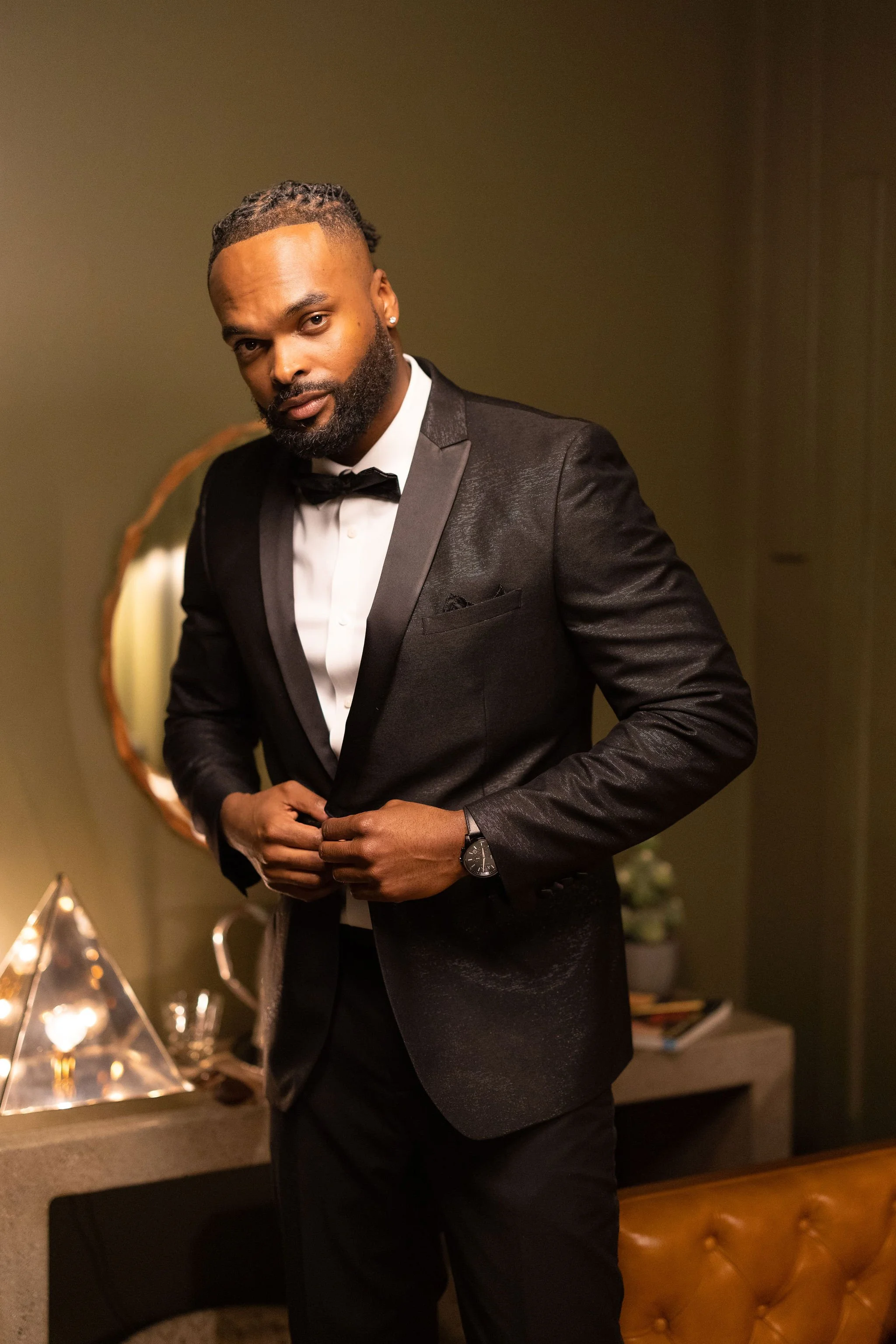 A man dressed in a black tuxedo with a bowtie, adjusting his suit, stands in an elegant room with warm lighting, a mirror, and decorative elements.