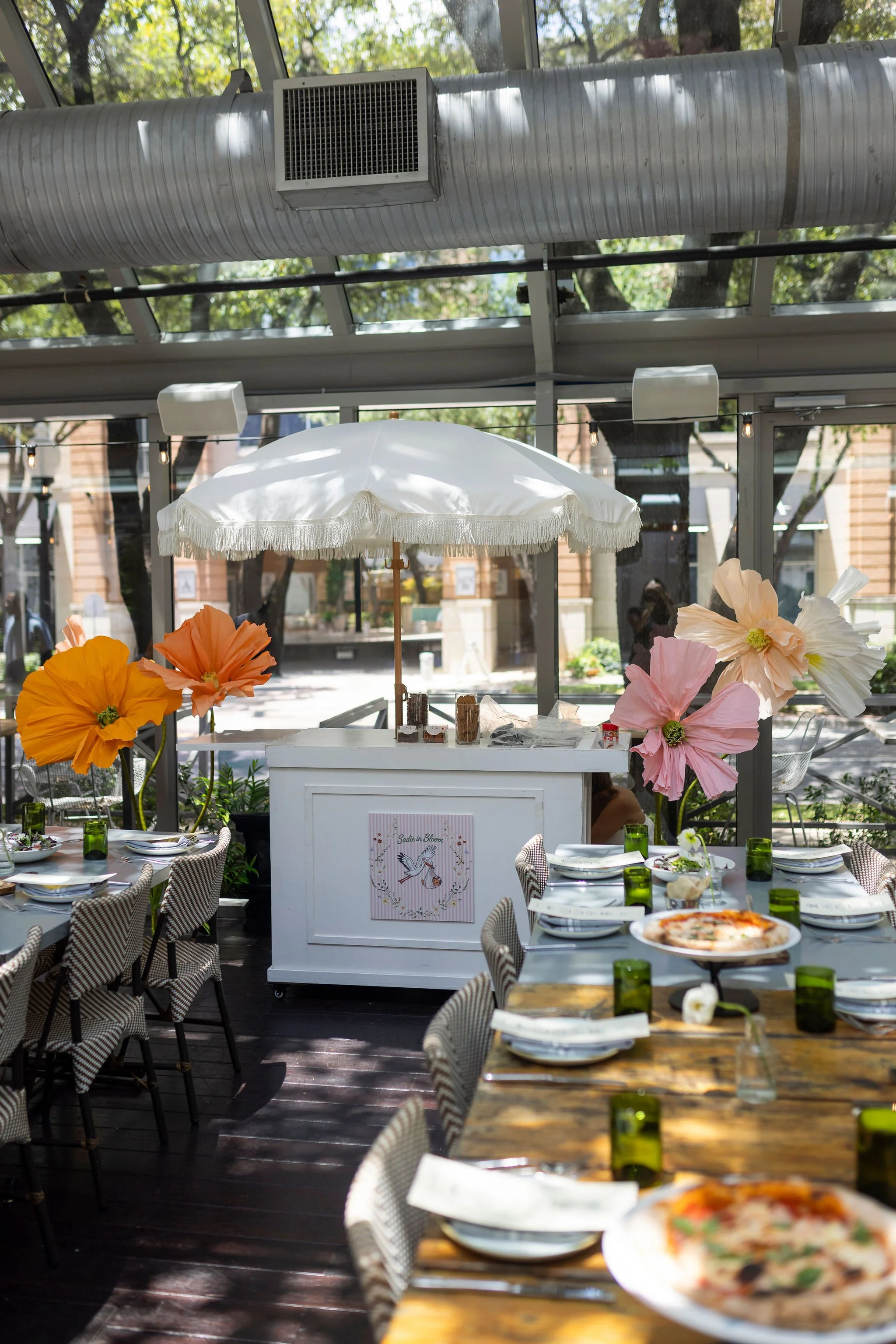 A restaurant or café with tables set for dining, decorated with large paper flowers, under a glass ceiling with trees visible outside, and a white wafer umbrella in the center.