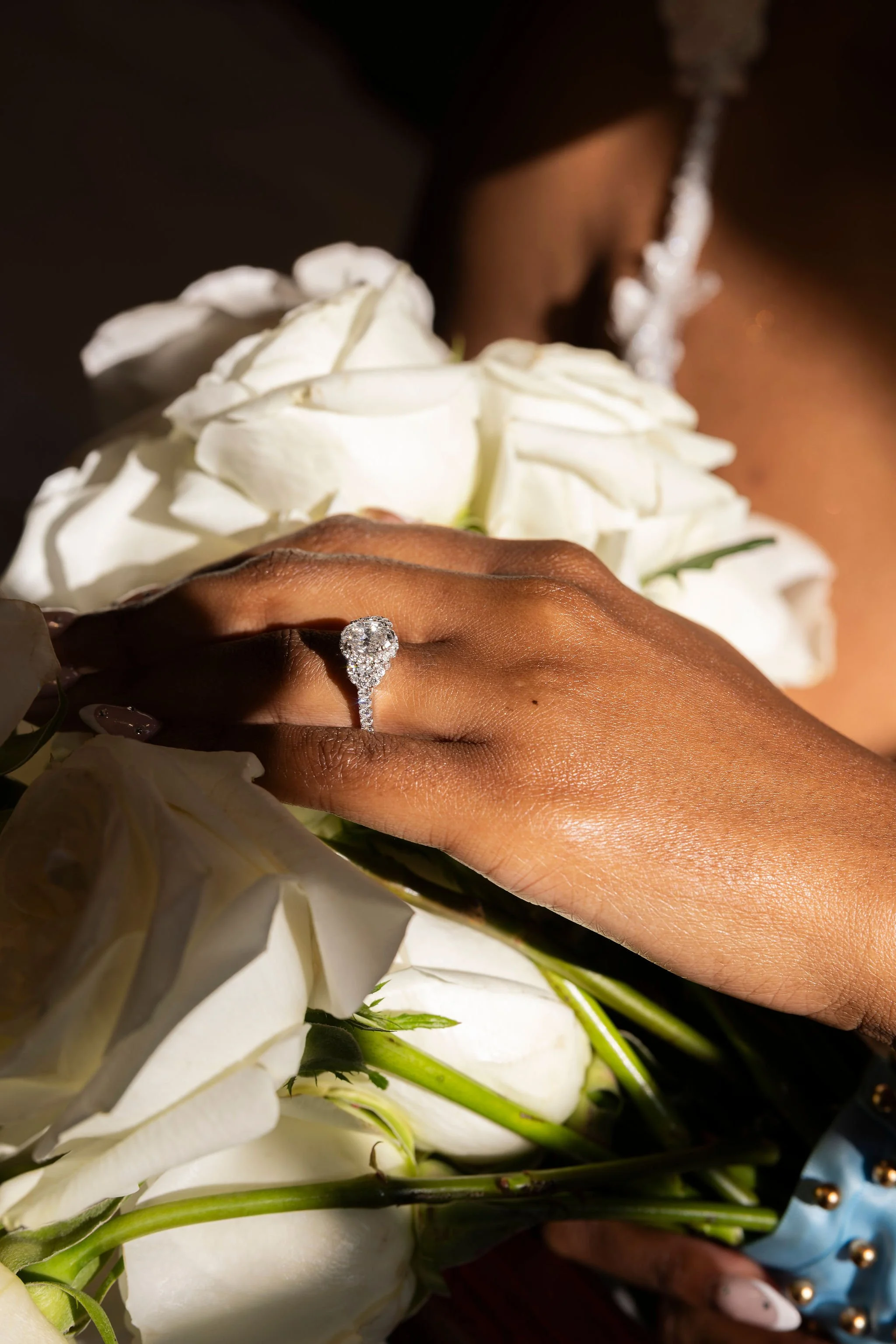 Close-up of a woman wearing a ring with diamonds, holding a bouquet of white and cream-colored flowers, with a blurred background.