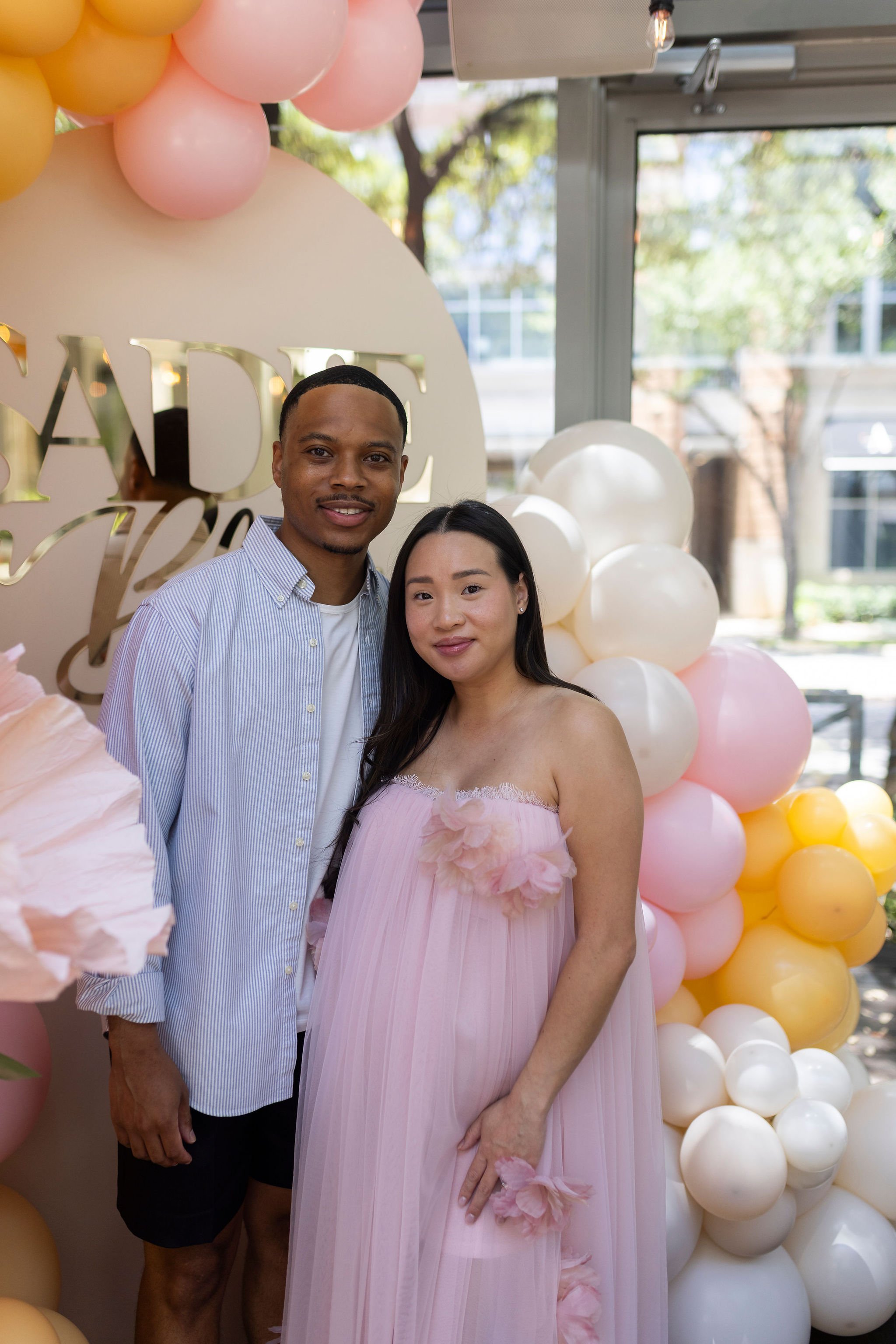 A couple standing in front of a balloon decoration with pink, white, and yellow balloons, and a sign that reads 'BABY' in gold letters. The man is wearing a light blue striped shirt and shorts, and the woman is wearing a pink strapless dress with flo