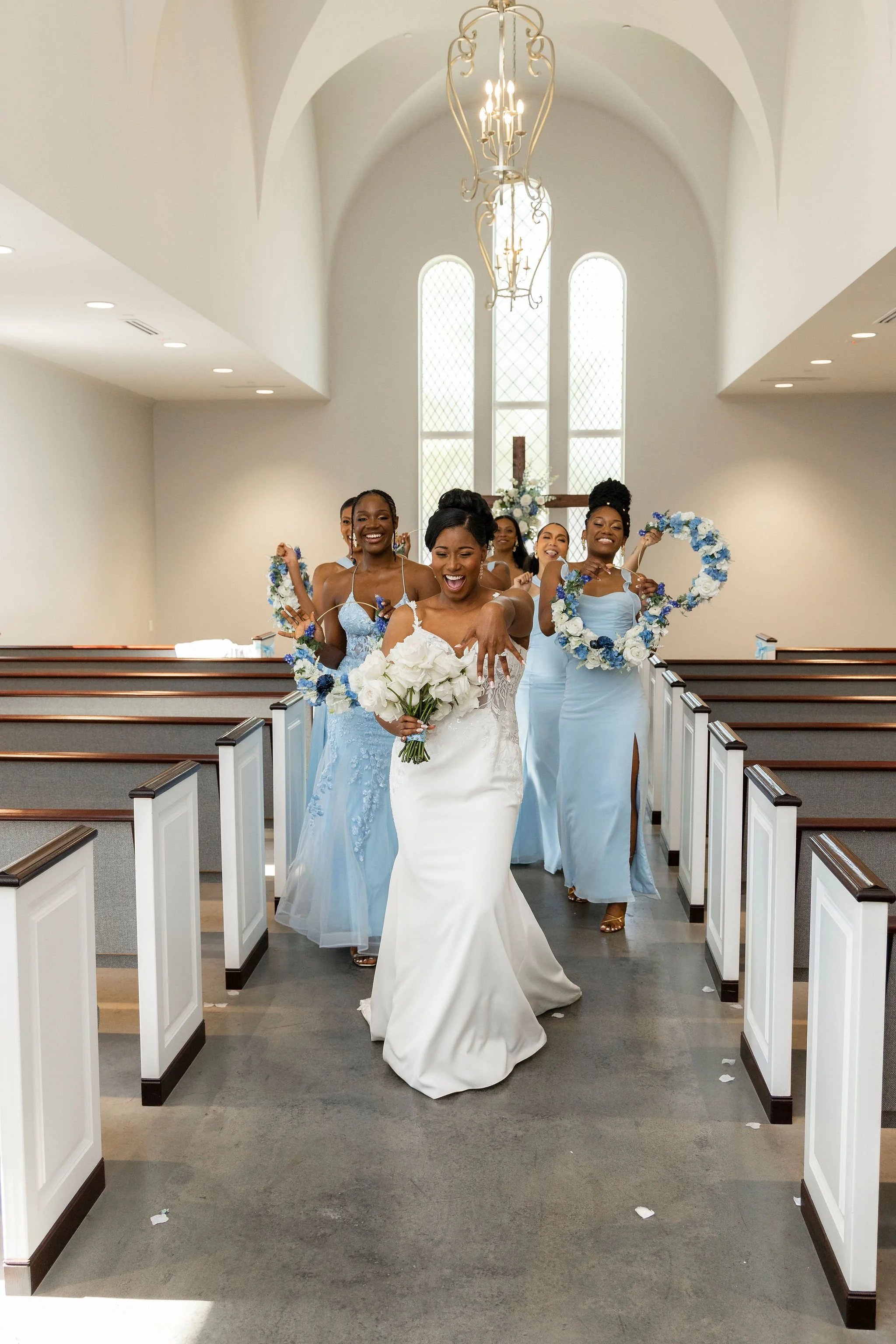 Bridal procession of women exiting a church, dressed in light blue and white gowns, carrying flowers and flower wreaths, with a cross and stained glass windows in the background.