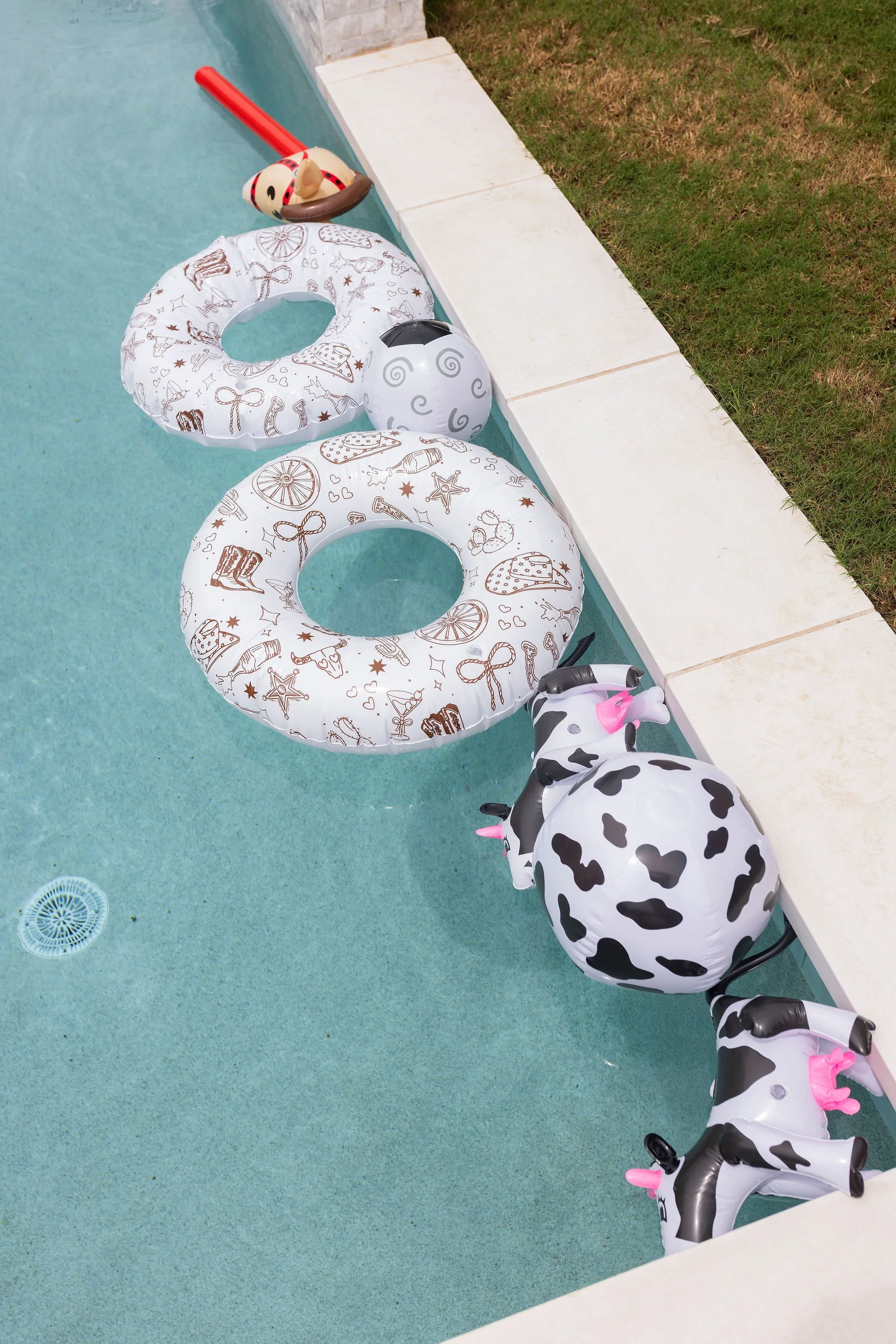 Inflatable pool floats including two white floats with chocolate and caramel dessert patterns, a black and white swirl float, and a cow-shaped float with pink accents, all on a light blue pool deck next to grass.