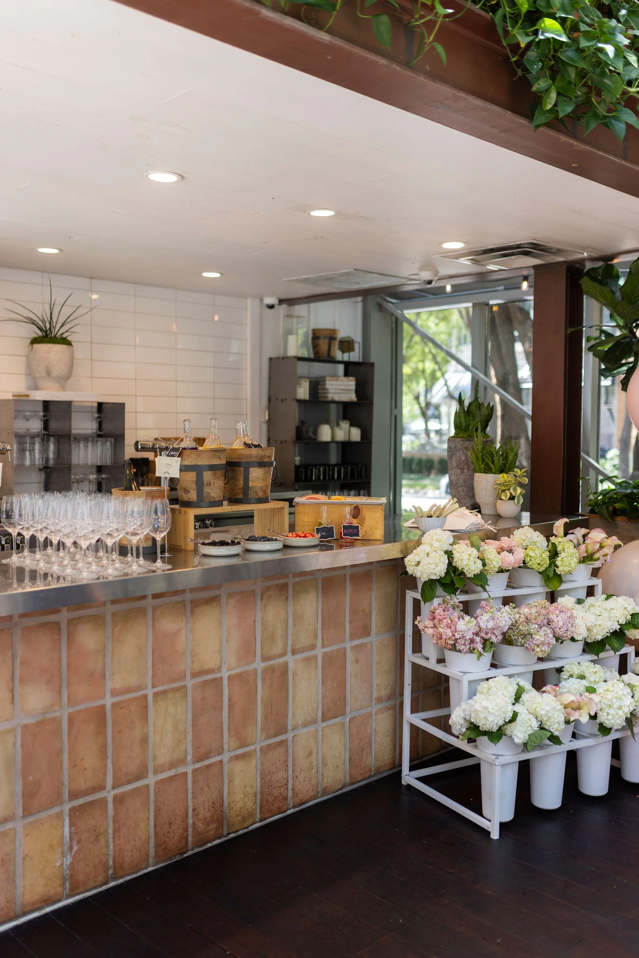Interior of a cafe or flower shop with a counter made of brick and a display of white and pink hydrangeas in white pots. There are glass bottles, small bowls of berries, and a shelf with glasses and other items in the background.