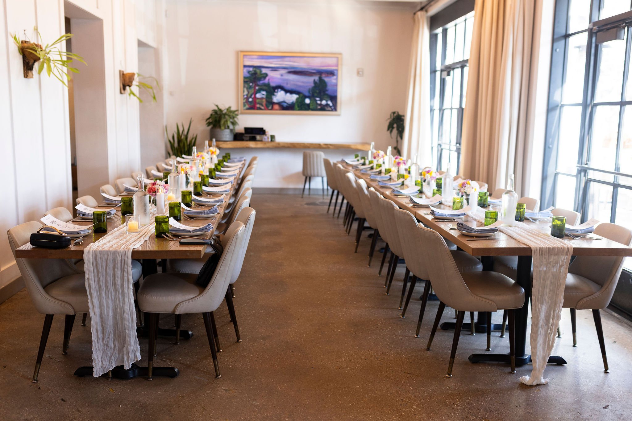 A long table set for a formal event in a bright room with large windows, plants, a painting on the wall, and place settings with plates, folded napkins, glasses, and small flower arrangements.