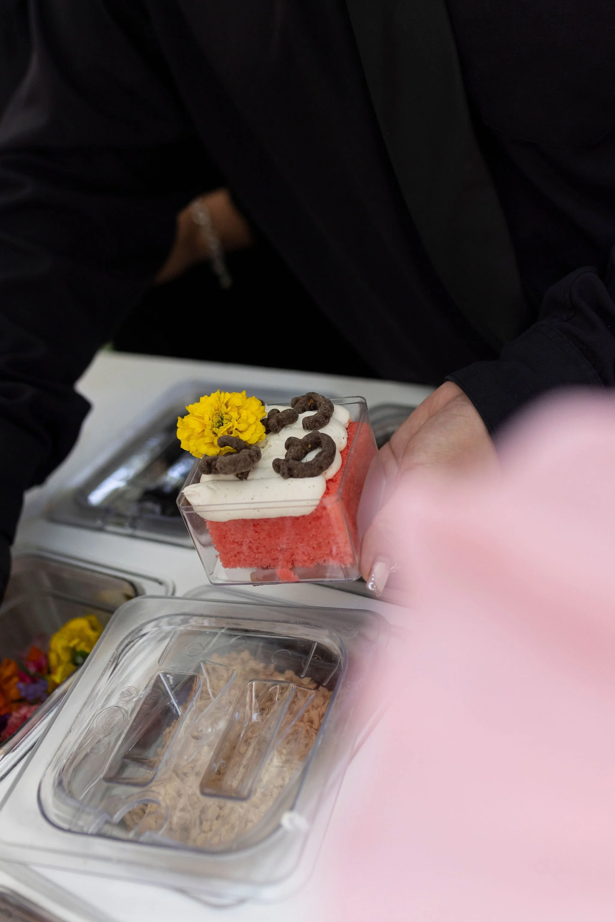 A small slice of pink cake with white frosting, chocolate pieces, and a yellow flower on top, held by a person in a dark outfit.