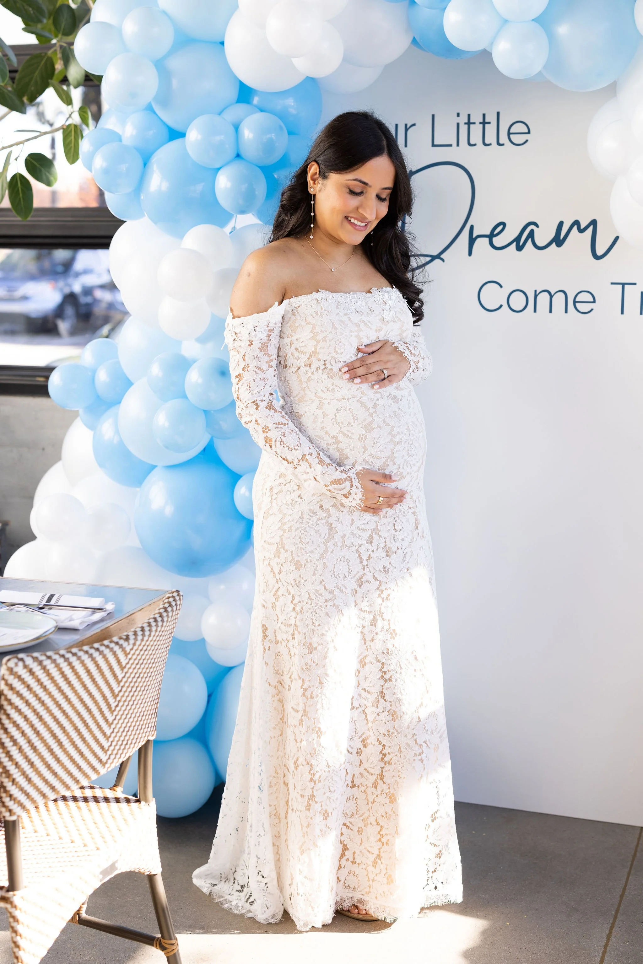 A pregnant woman in a white lace dress stands with one hand on her belly at a baby shower decorated with blue and white balloons and a sign that reads "Your Little Dream Come True."