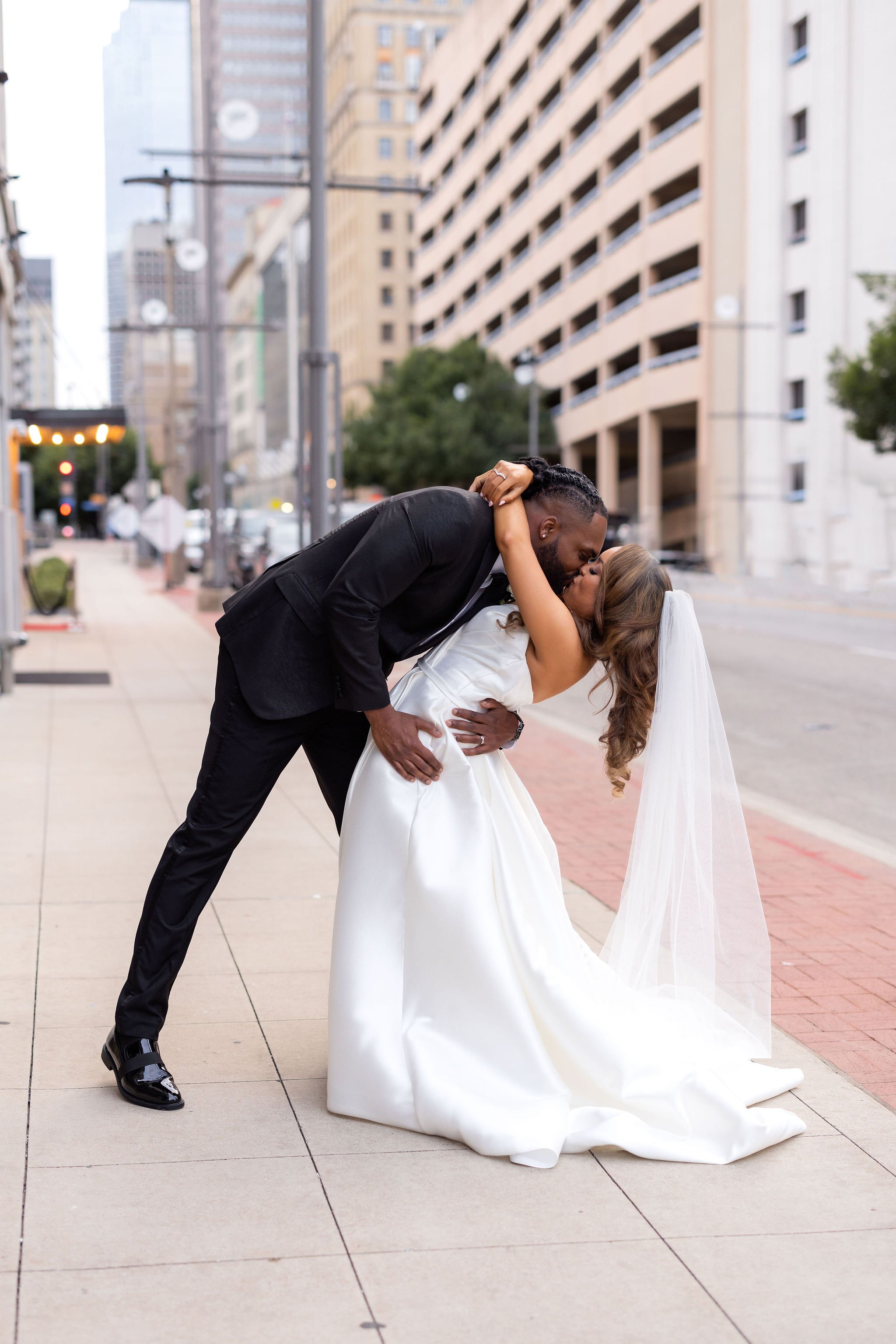 A joyful couple in wedding attire sharing a romantic kiss on a city sidewalk, with tall buildings in the background, during daytime.